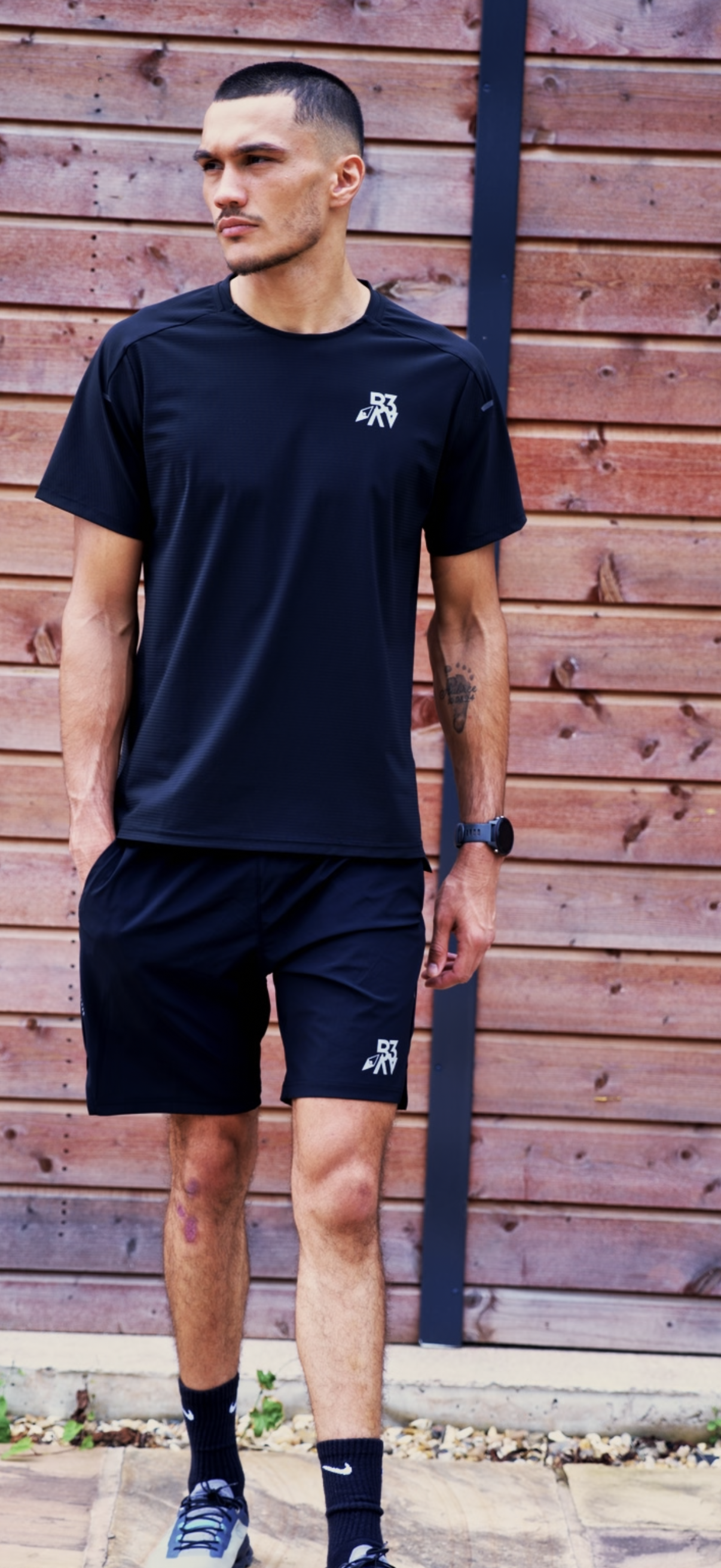 A man standing outdoors in athletic clothing, including a black t-shirt and shorts, with a wooden fence background.