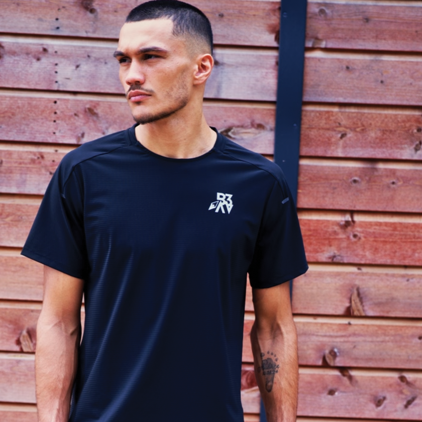 Young man with short hair and a slight beard stands outside in front of a wooden fence, wearing a black athletic shirt with a small logo on the chest.