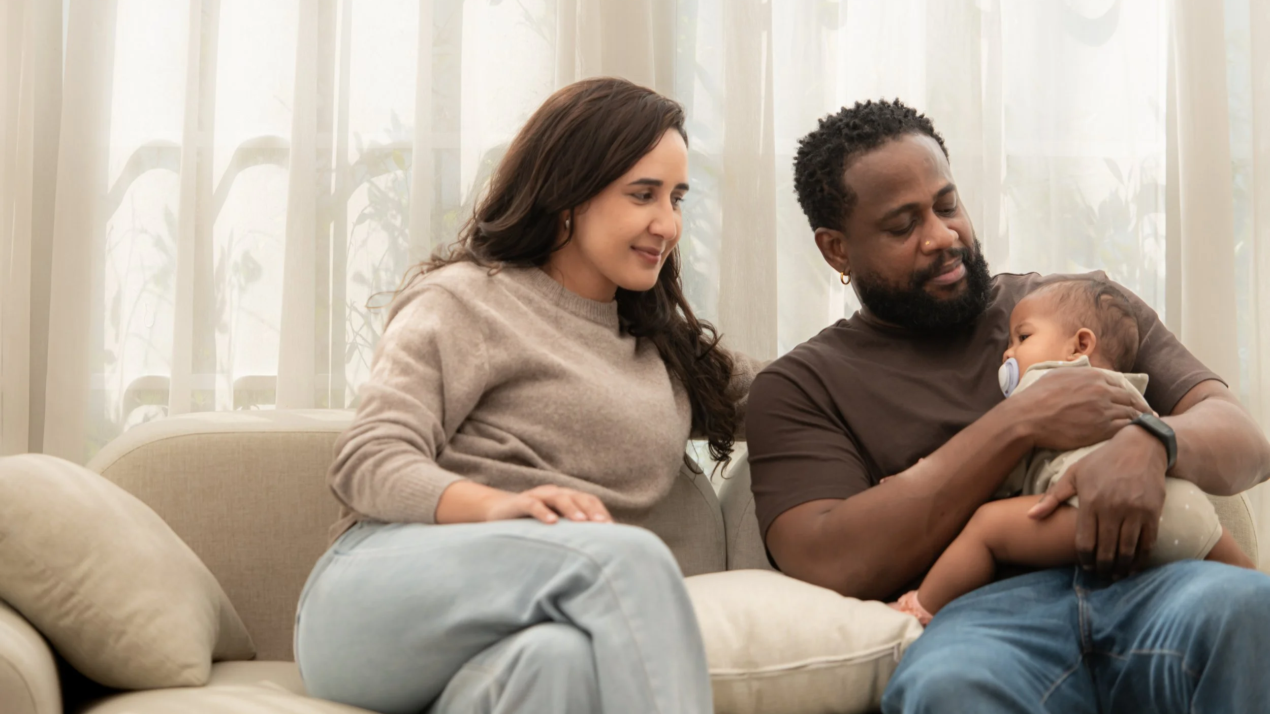 A family sitting on a beige sofa in a living room. The mother has long brown hair and is wearing a beige sweater. The father has a beard, wears a dark T-shirt, and is holding a baby with a pacifier. The background features light-colored curtains and plants outside the window.