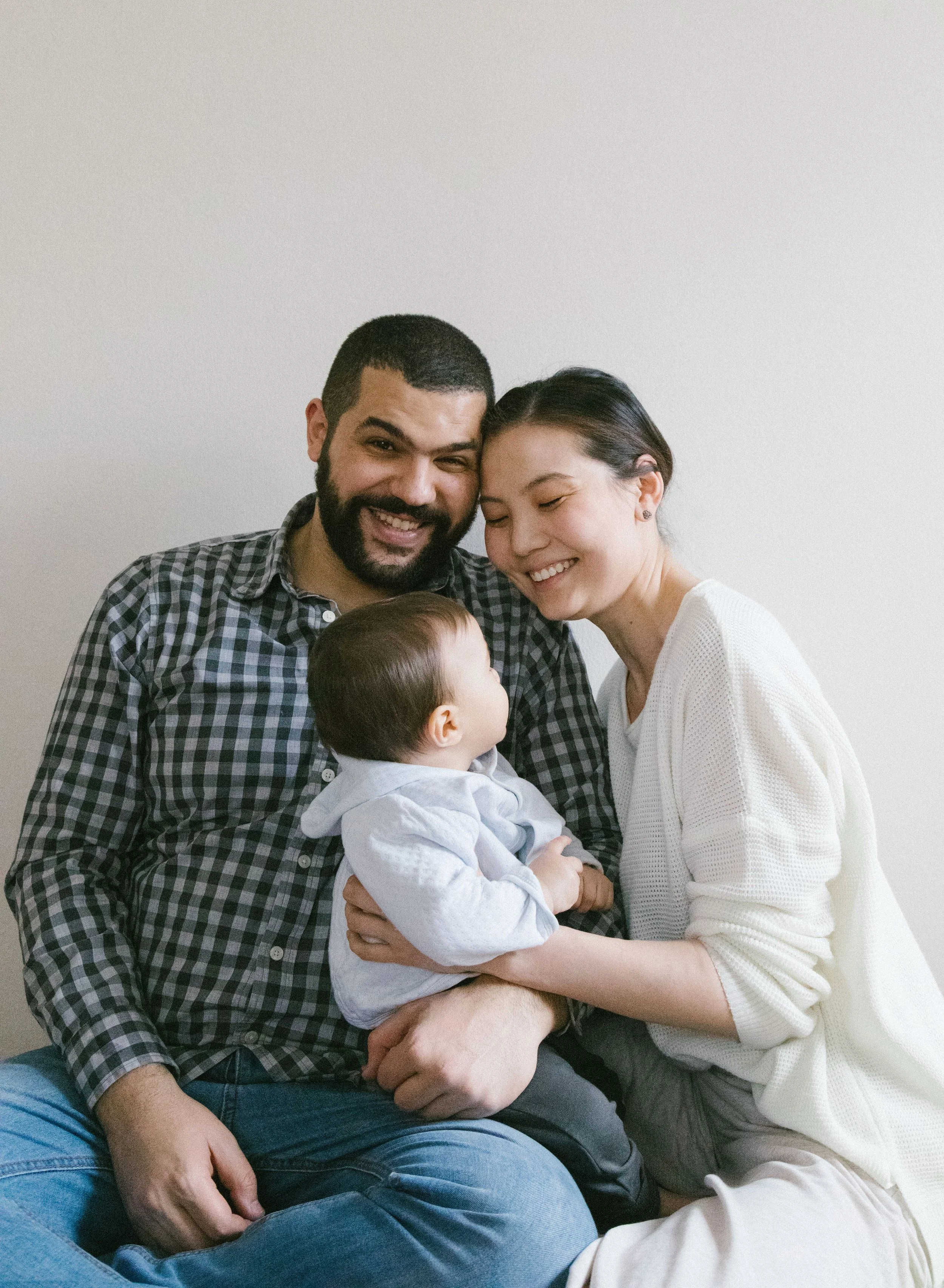A happy multicultural family of three including a man, a woman, and a young child, sitting close together and smiling against a plain white background.