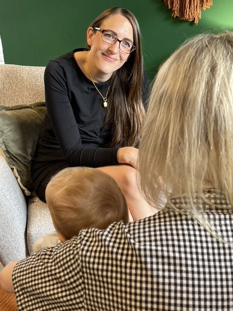 A woman with long brown hair and glasses sitting on a beige couch, smiling at the camera, while a blonde-haired adult and a young child sit in front of her, facing away.