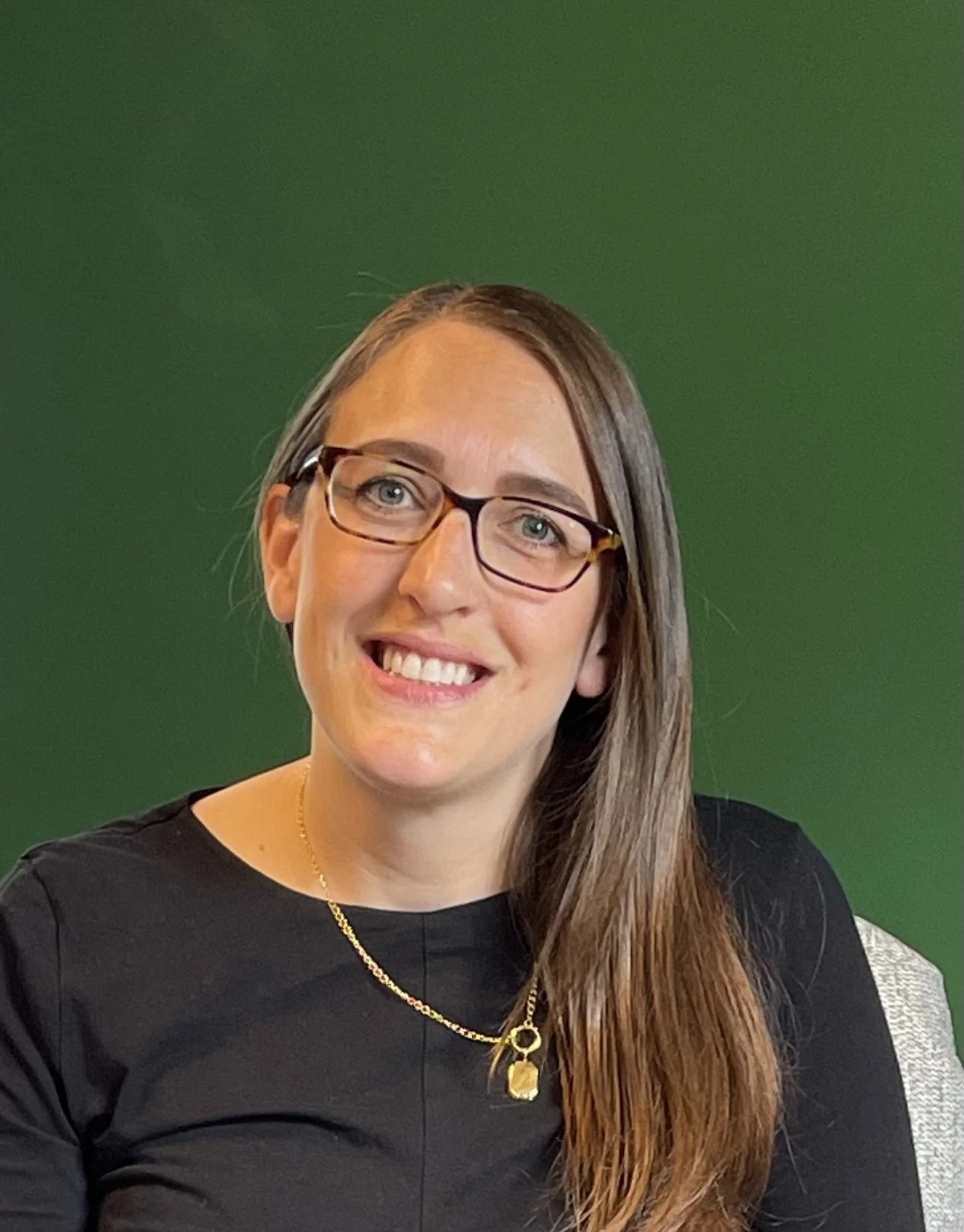 A woman with glasses and long brown hair smiling, wearing a black shirt and a gold necklace, sitting against a green background.