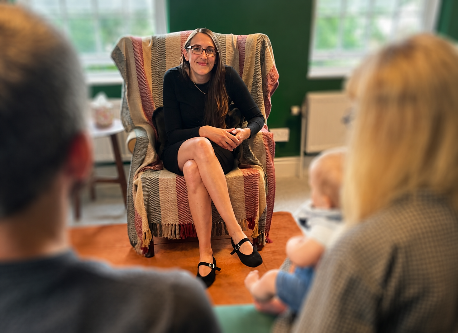 Woman sitting in a patterned armchair, speaking to a small group in a cozy room with green walls and large windows.