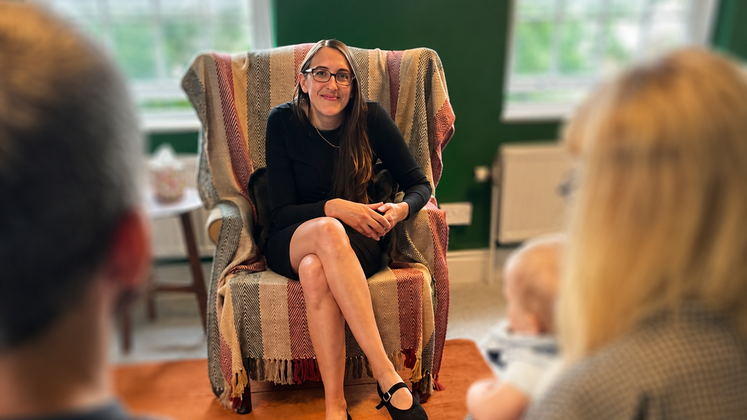 A woman with long brown hair, glasses, wearing a black dress, sitting in a patterned armchair with a colorful throw blanket, speaking to two adults and a baby in a cozy room with green walls and windows in the background.