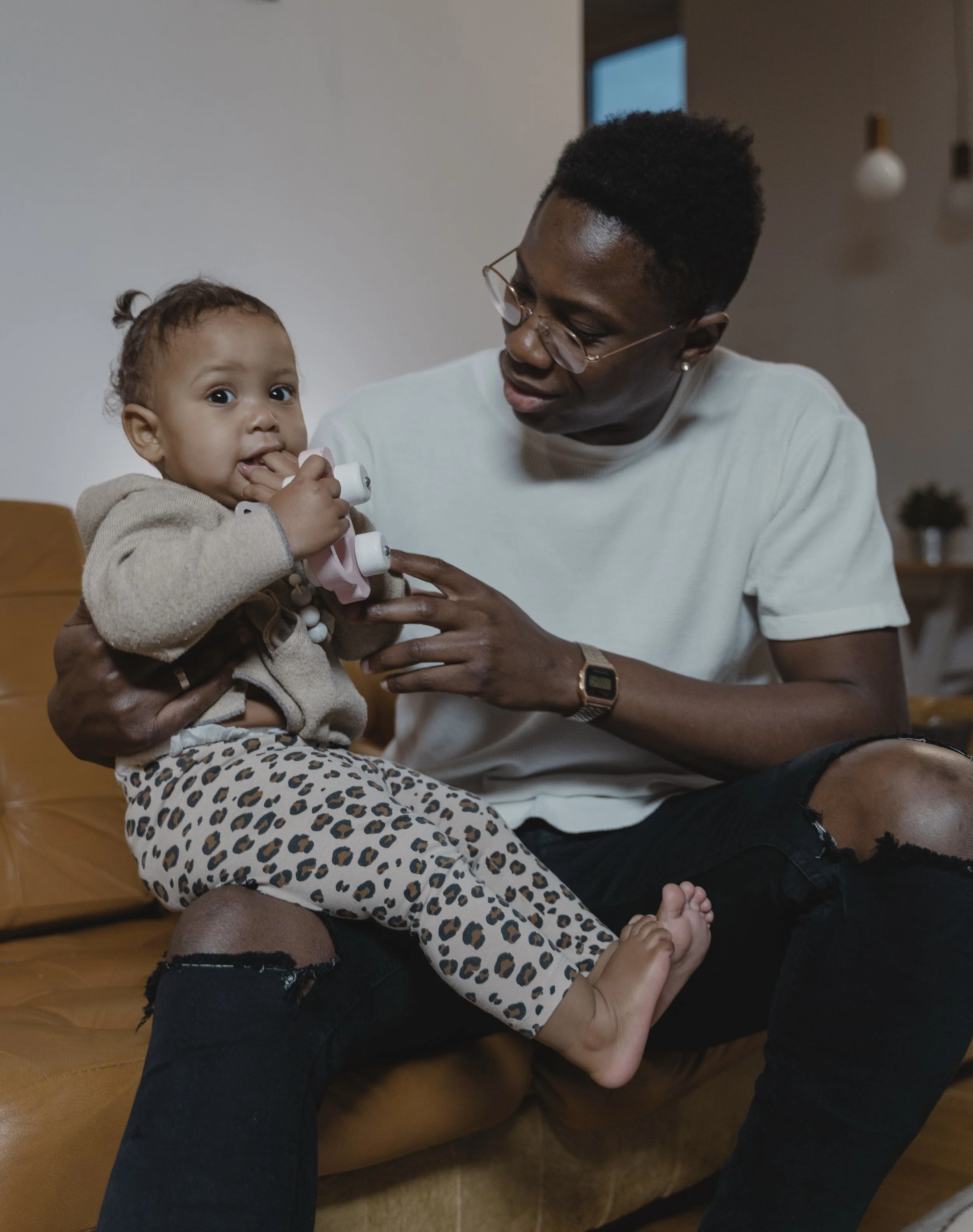 A woman holding a young child sitting on a leather sofa in a living room, with her lap visible. The child is looking at the camera, holding a toy phone, and wearing leopard print pants and a beige jacket.
