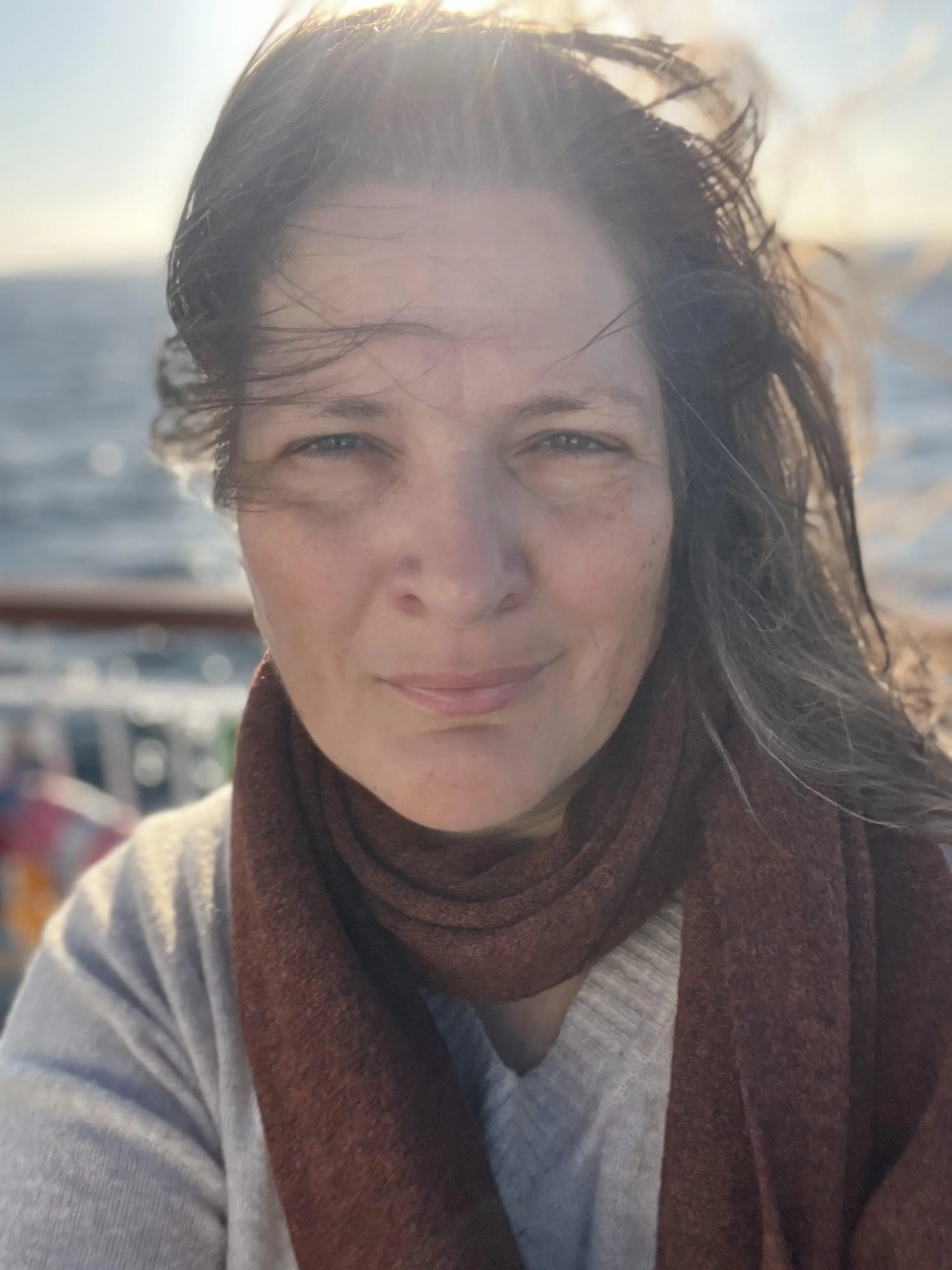 Close-up of a smiling woman with windblown hair against a background of the ocean and sky during sunset.