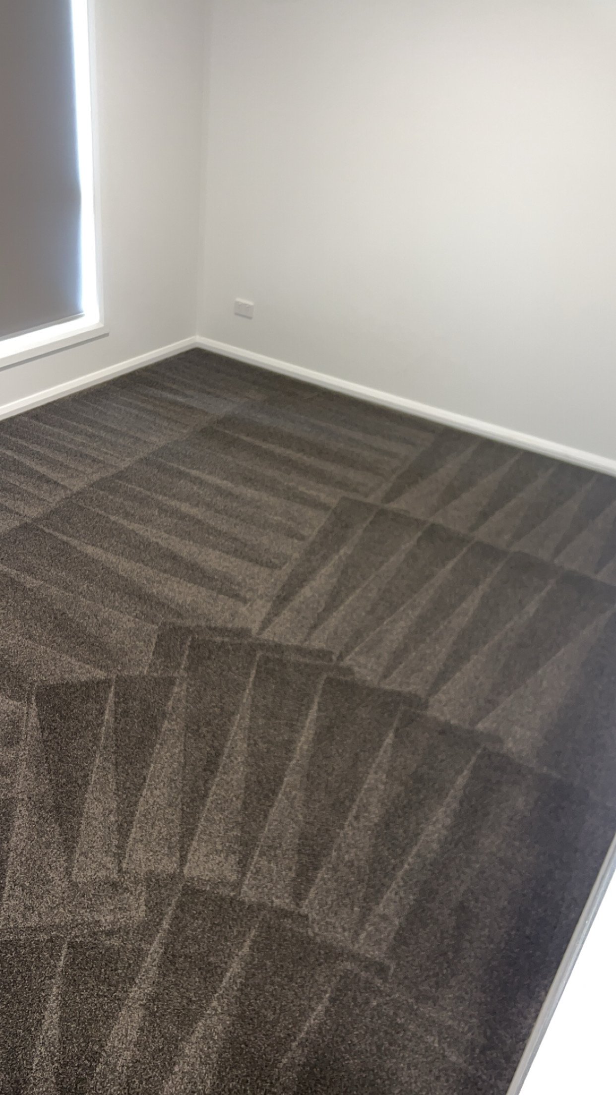Empty room with white walls, a window with a closed gray blind, electrical outlets, and carpet flooring with geometric patterns.