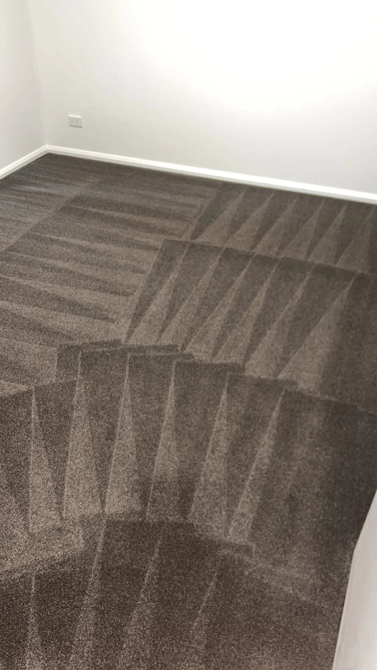 Carpeted floor with a geometric pattern in shades of brown and tan, and plain white walls with a white baseboard and a power outlet.
