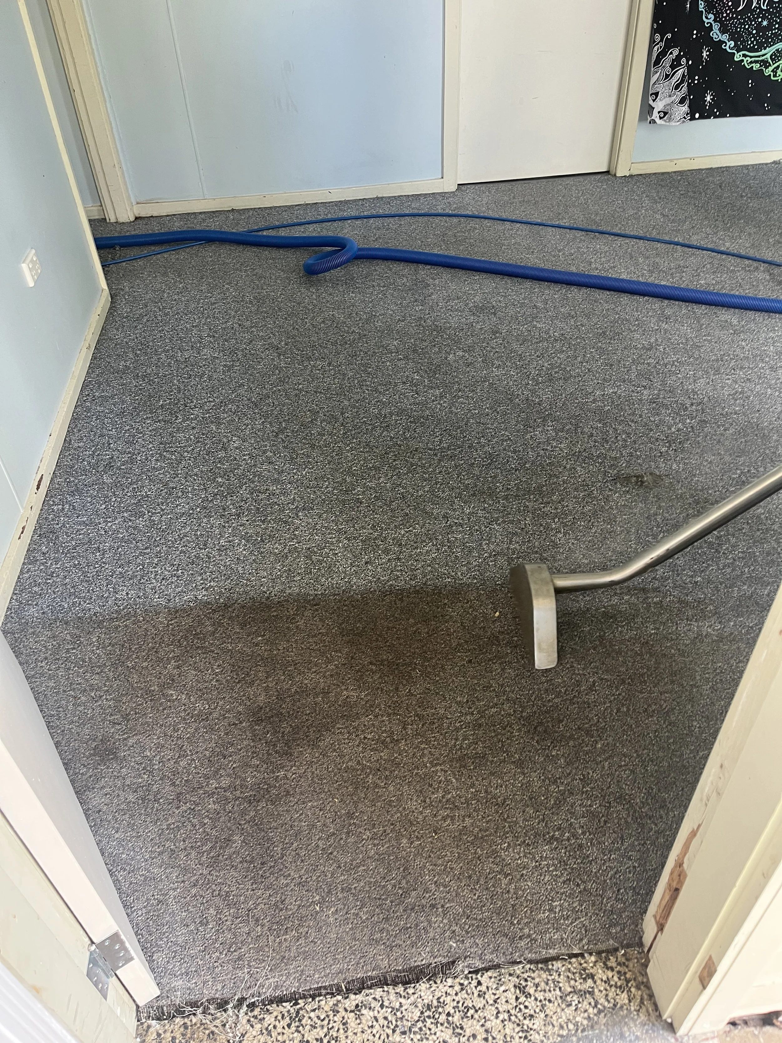 An open doorway leading from a room with a grey carpet to a room with a darker grey textured carpet. A blue hose is lying on the grey carpeted floor, coiled near the door. The door frame is white, and the walls are light blue.