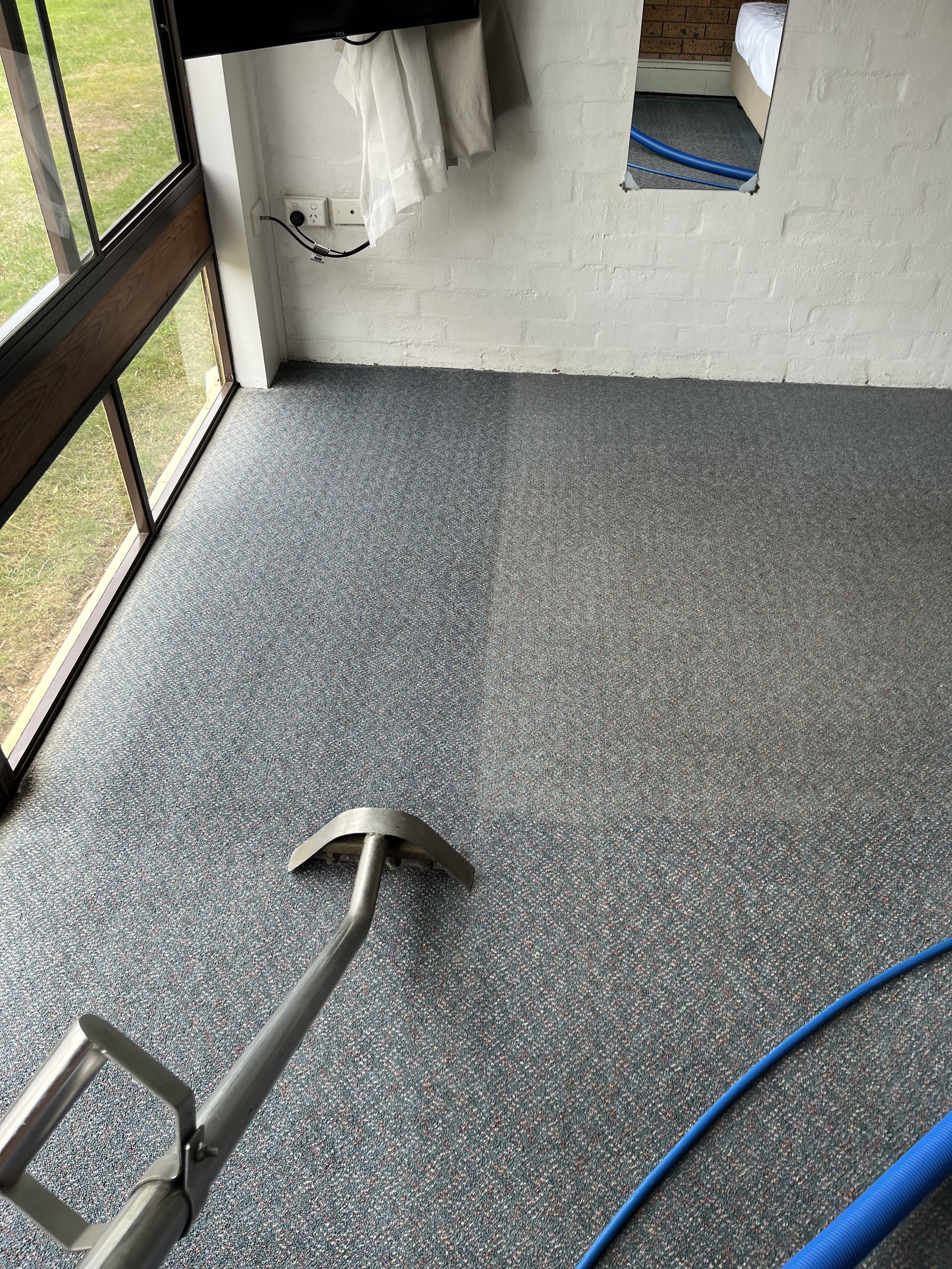 An empty room with a sliding glass door, a white brick wall, a partially visible bed, a blue hose on the carpet, and a small treadmill with a metal frame.