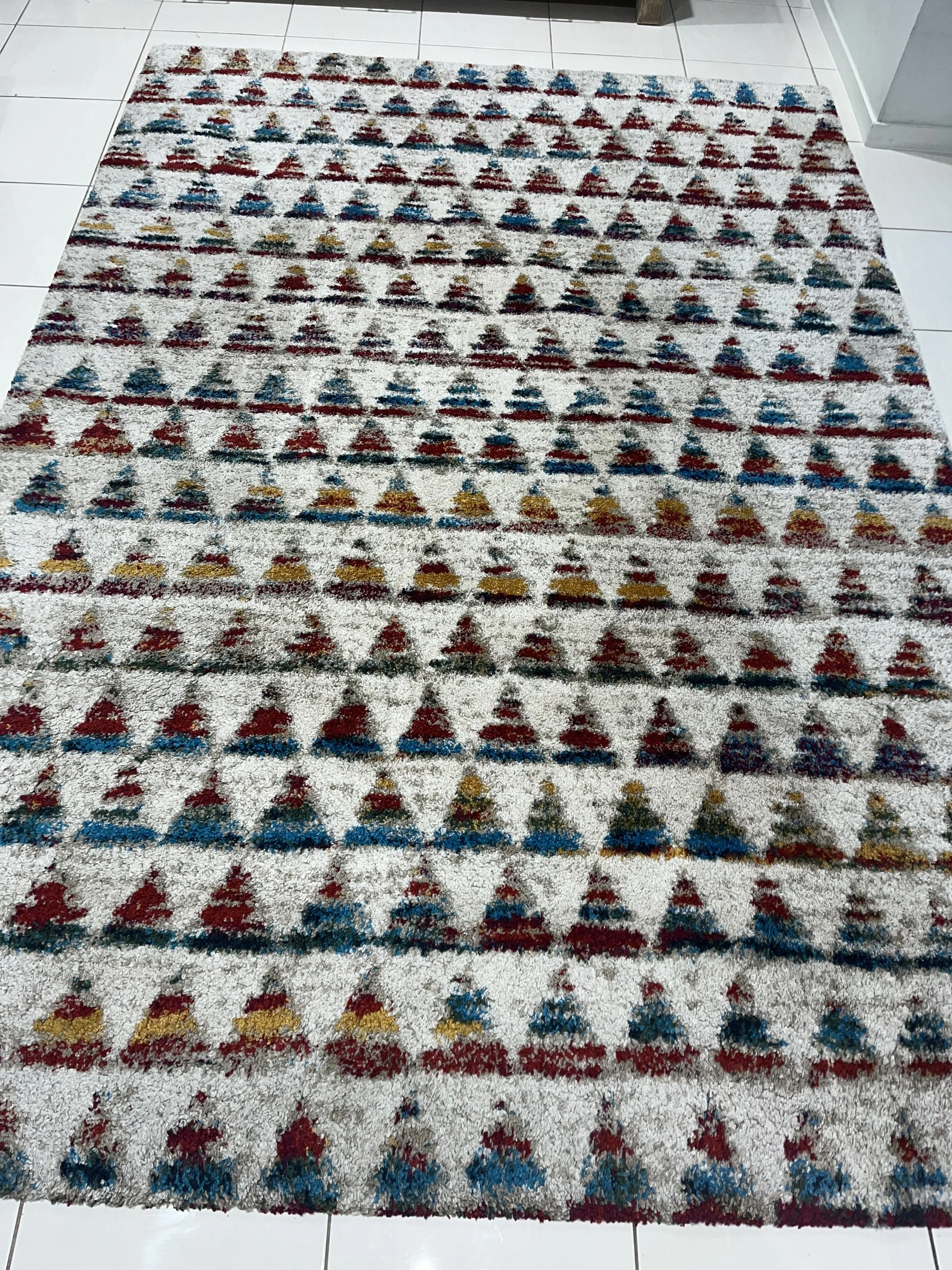 A rectangular rug with a geometric pattern of triangles in red, blue, yellow, and black on a white background, placed on a tiled floor.
