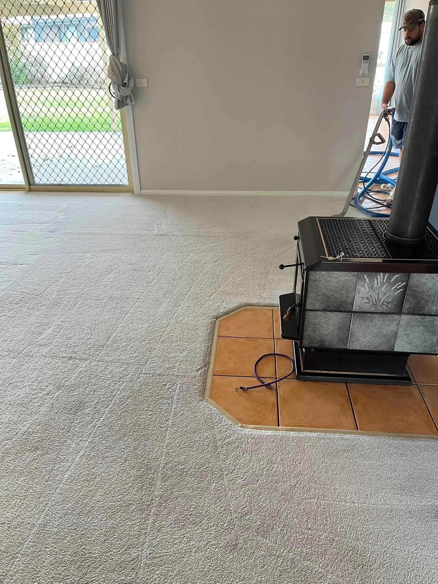 Interior of a living room with beige carpet, a sliding glass door with a dog gate, and a person vacuuming the floor near a black stove or fireplace. There's a small wooden landing or step with tiles.