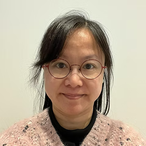 A woman with short dark hair wearing glasses, a black shirt, and a pink sweater posing against a neutral background.