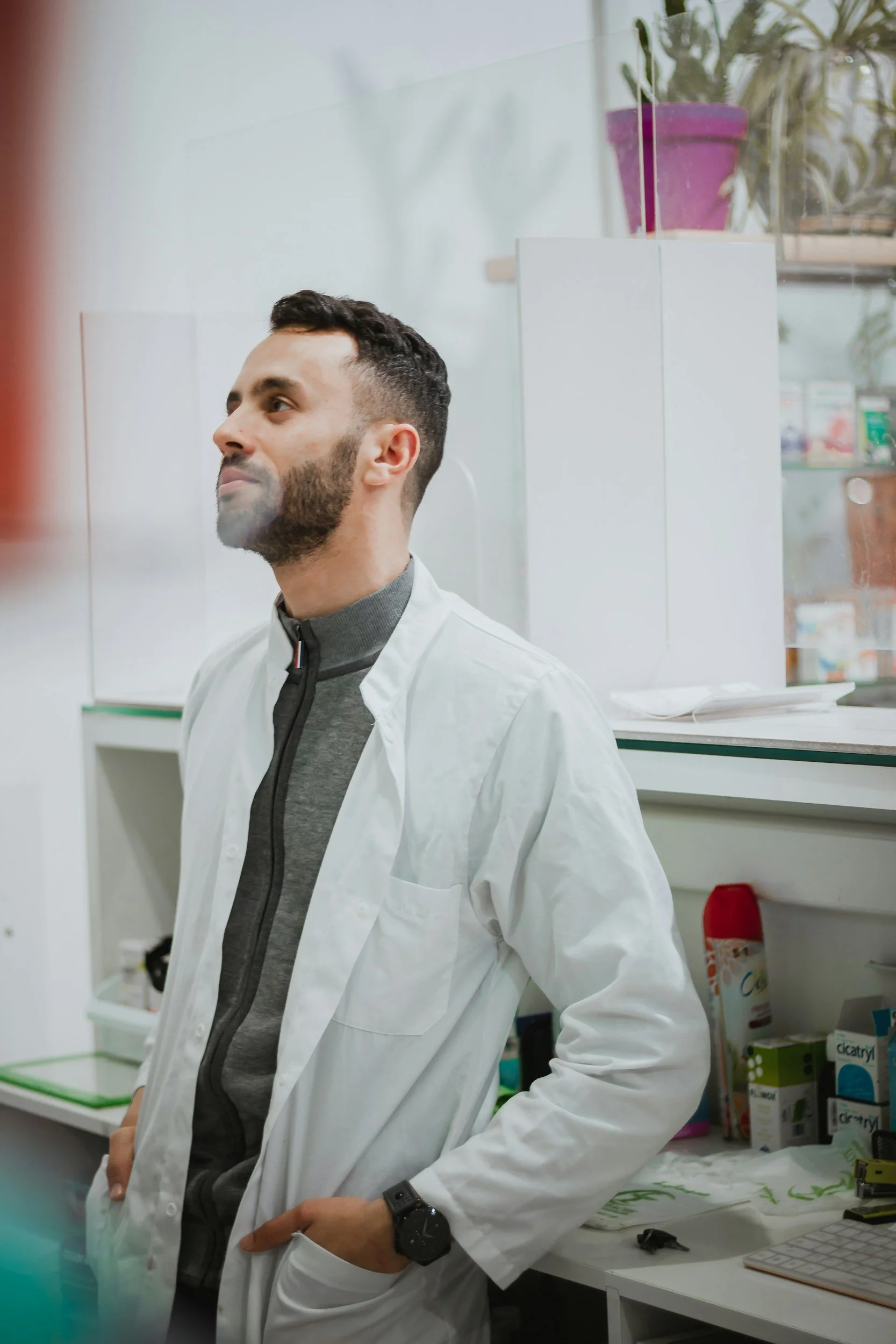 A man with dark hair and a beard wearing a white lab coat, a gray shirt, and a black watch, standing in a laboratory with various supplies and green plants in the background.