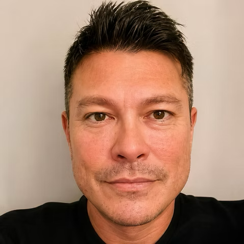 Close-up portrait of a man with short dark hair, light skin, and wearing a black shirt, looking directly at the camera against a plain light background.