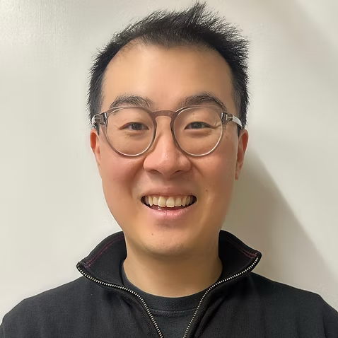 Portrait of a smiling man wearing glasses and a black zip-up jacket, standing against a plain white wall.