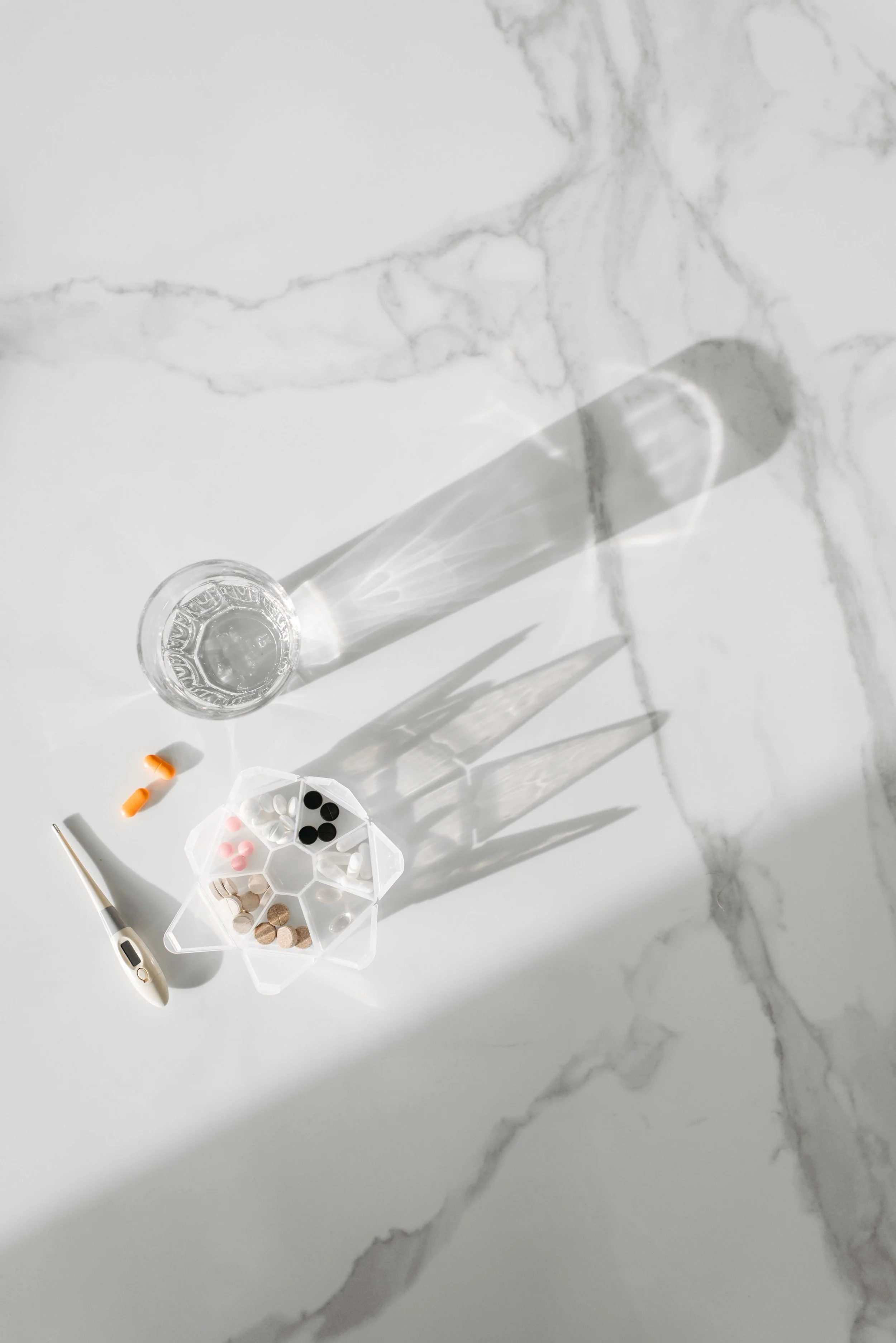 A marble surface with a glass of water, a thermometer, a pill organizer filled with various pills, a few loose pills, and a syringe.
