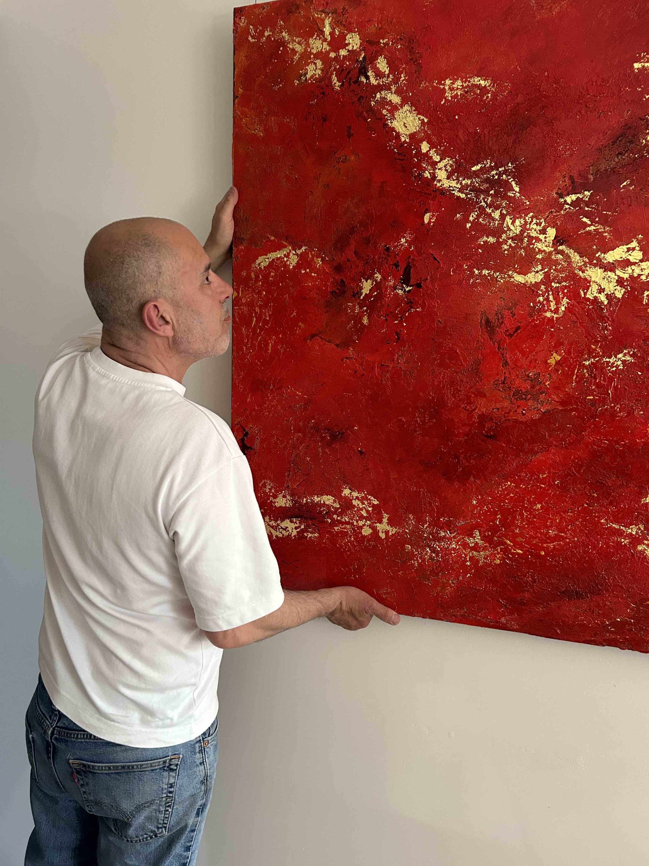 A man with a bald head and white t-shirt holding a large abstract red and gold painting against a plain wall.