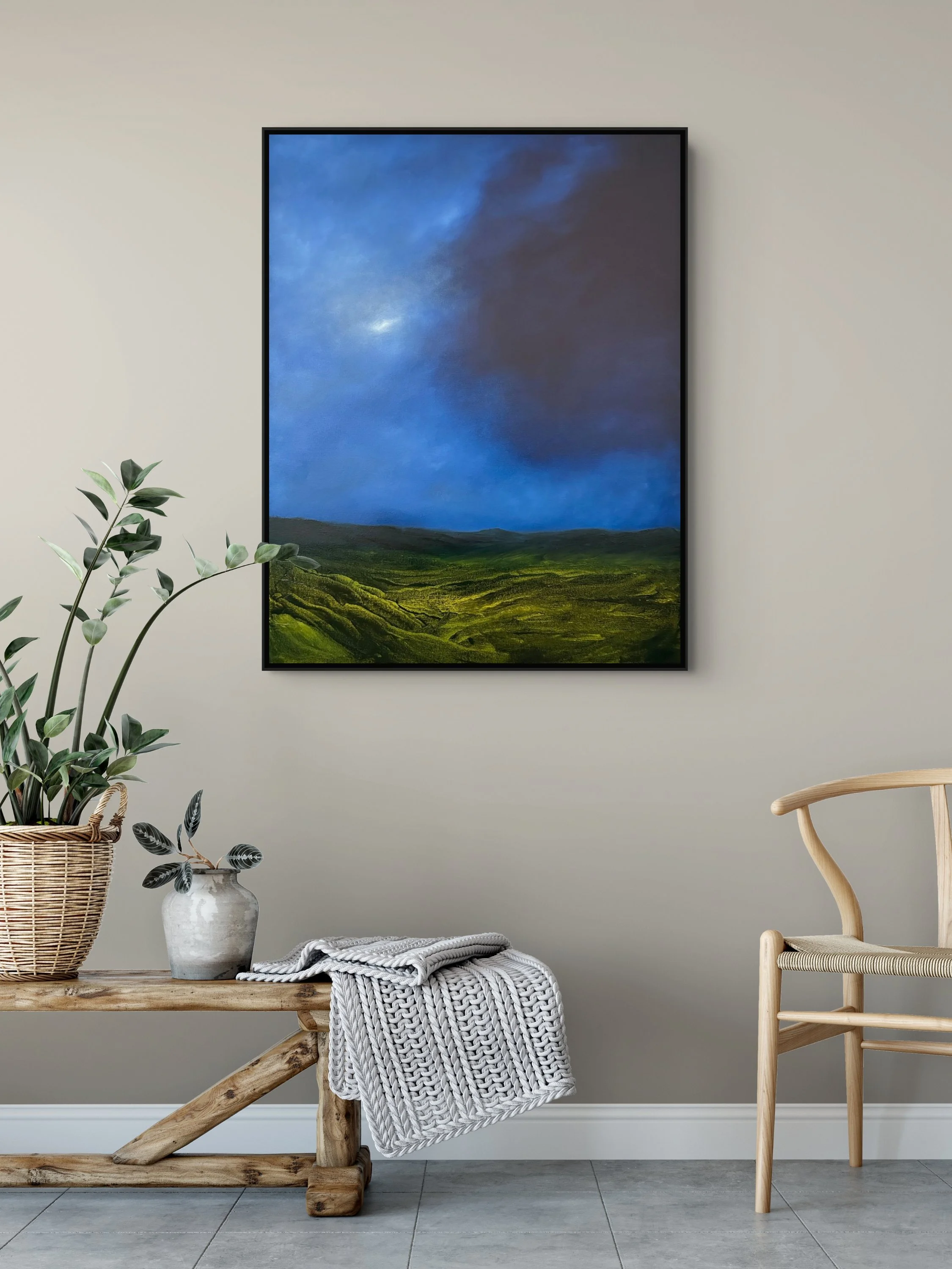 A minimalistic living space with a framed landscape painting of cloudy skies over green rolling hills hanging on a beige wall. On a rustic wooden table in the foreground, there are potted plants and a neatly folded gray and white knitted blanket. To the right, there's a light wooden chair with a woven seat.