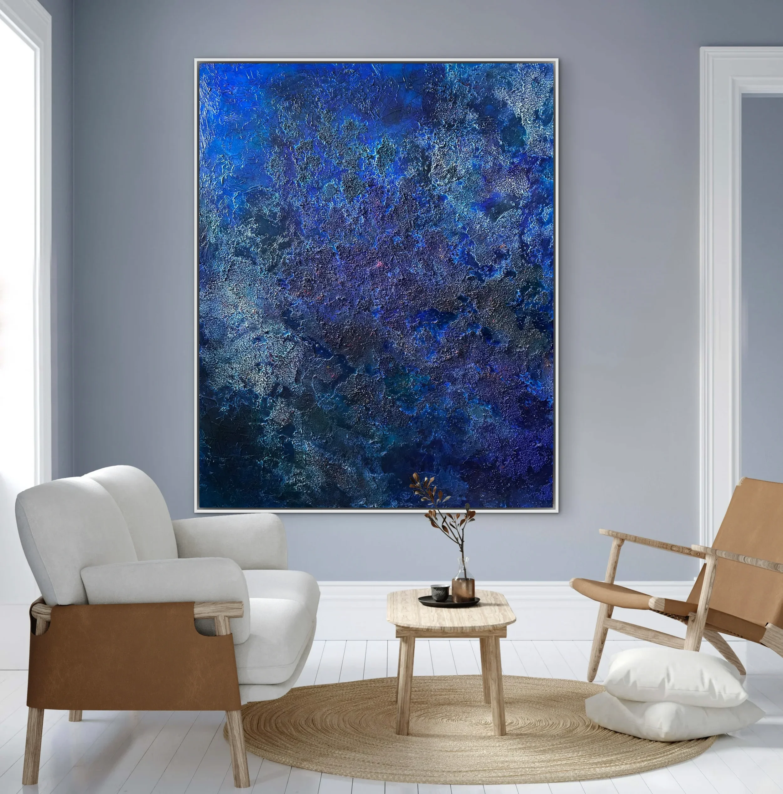 A modern living room with a large blue abstract painting on a gray wall, a white sofa, a small wooden coffee table with black accessories, a rattan rug, a leafy plant in a copper pot, and a wooden chair with a woven seat.