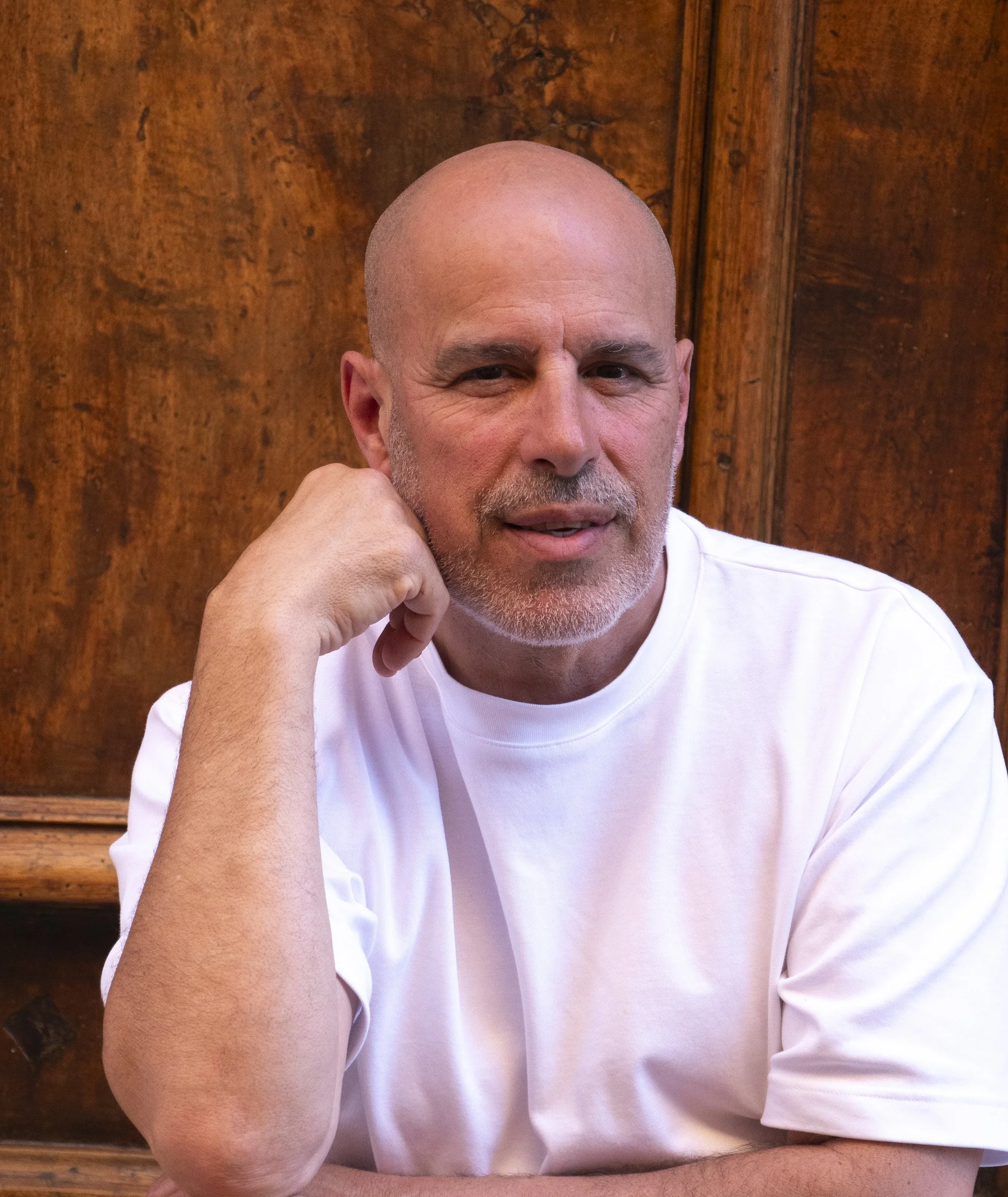 A middle-aged bald man with a beard is sitting by a wooden wall, resting his chin on his hand and looking at the camera.