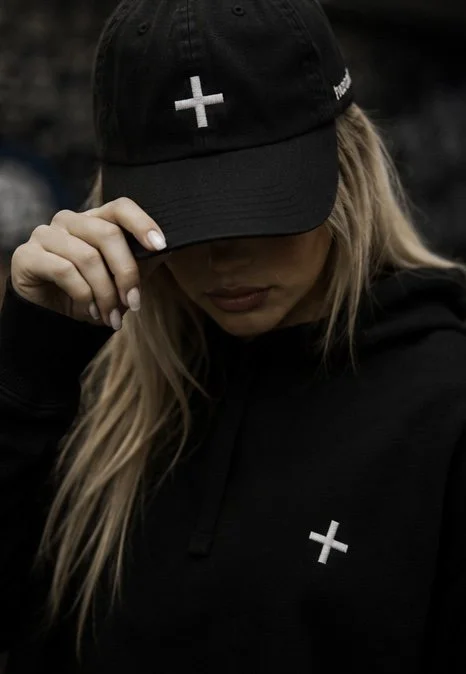 A woman wearing a black baseball cap and hoodie, both with white plus signs.
