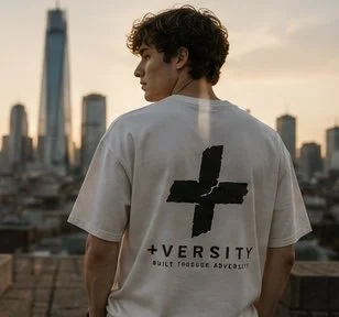 Young man with curly hair wearing a white T-shirt with a black cross and the text '+VERSITY' on the back, city skyline at sunset in the background.