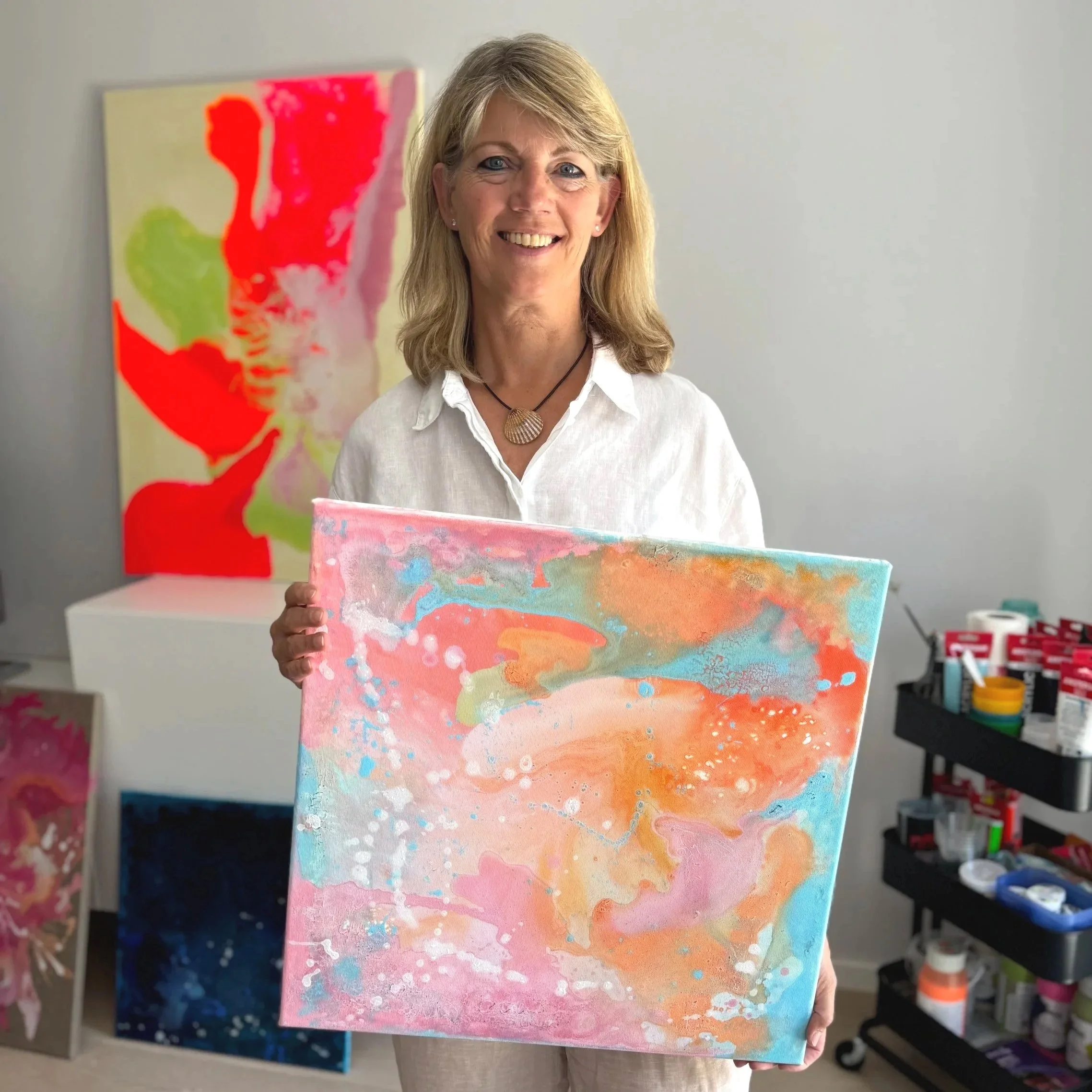 Eine Frau mit blonden Haaren hält ein buntes abstraktes Gemälde in den Händen. Im Hintergrund sind weitere Kunstwerke und Kunstbedarf sichtbar.