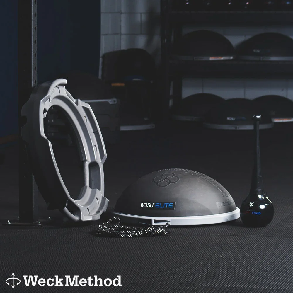 Fitness equipment including a circular support frame, BOSU Elite balance trainer, jogging or speed ladder, and a golf club-shaped equipment, arranged on a black gym floor with shelves of balance disks in the background.