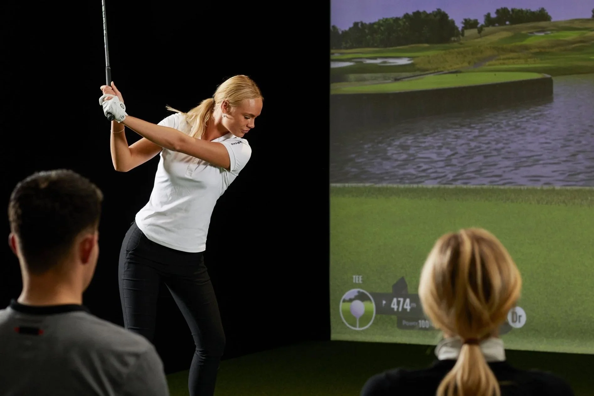 A woman in white polo shirt and black pants demonstrating a golf swing to a small group of people in front of a large screen showing a golf course landscape with water and greenery.