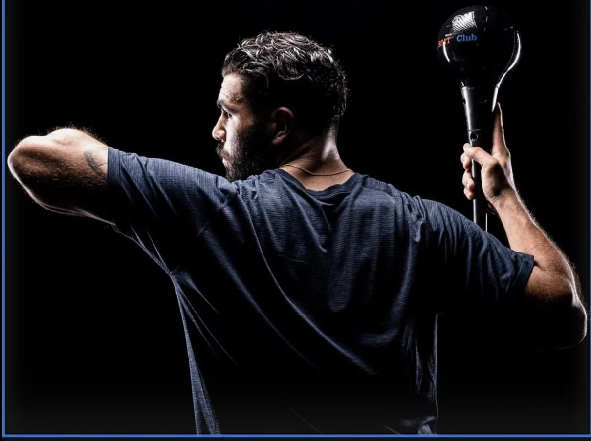 Man with dark hair and beard, wearing a navy athletic shirt, holding a microphone in his left hand, turned sideways, against a black background.