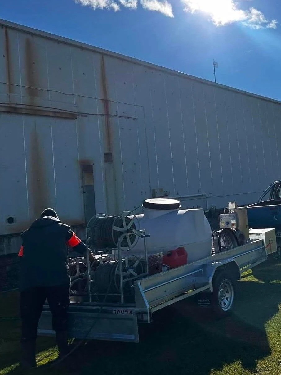 A person is refilling a building wash trailer attached to a truck, outside a large industrial building.