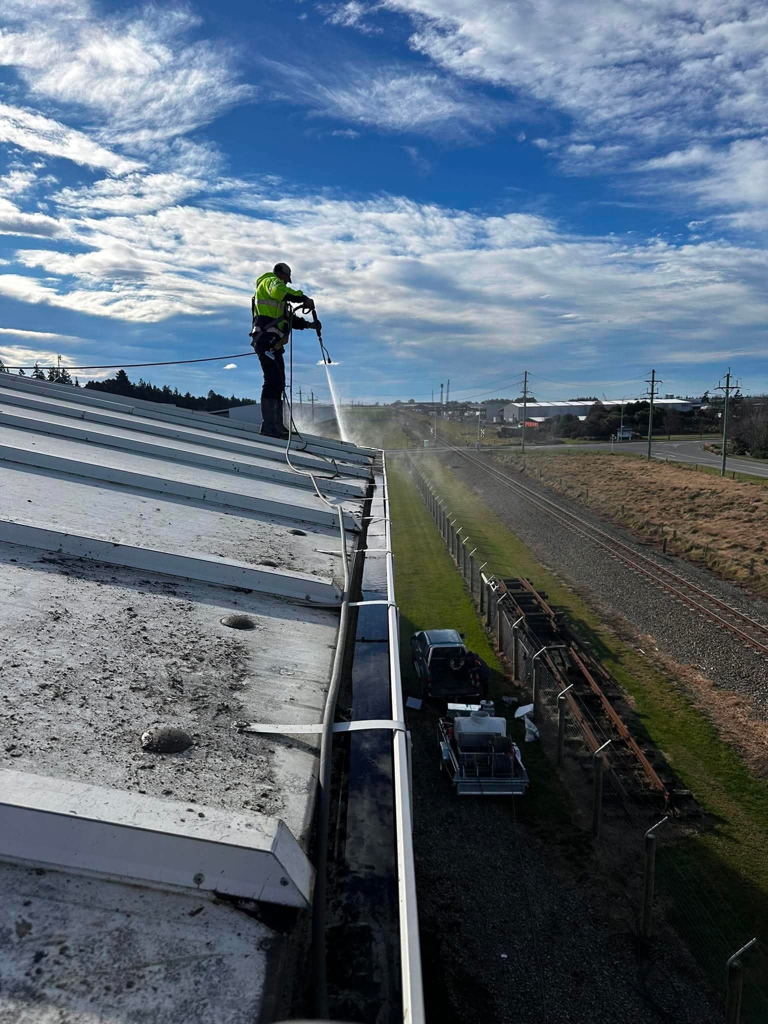 A worker in safety gear pressure washing the roof of a building, with a sky filled with clouds above and train tracks below in the background.