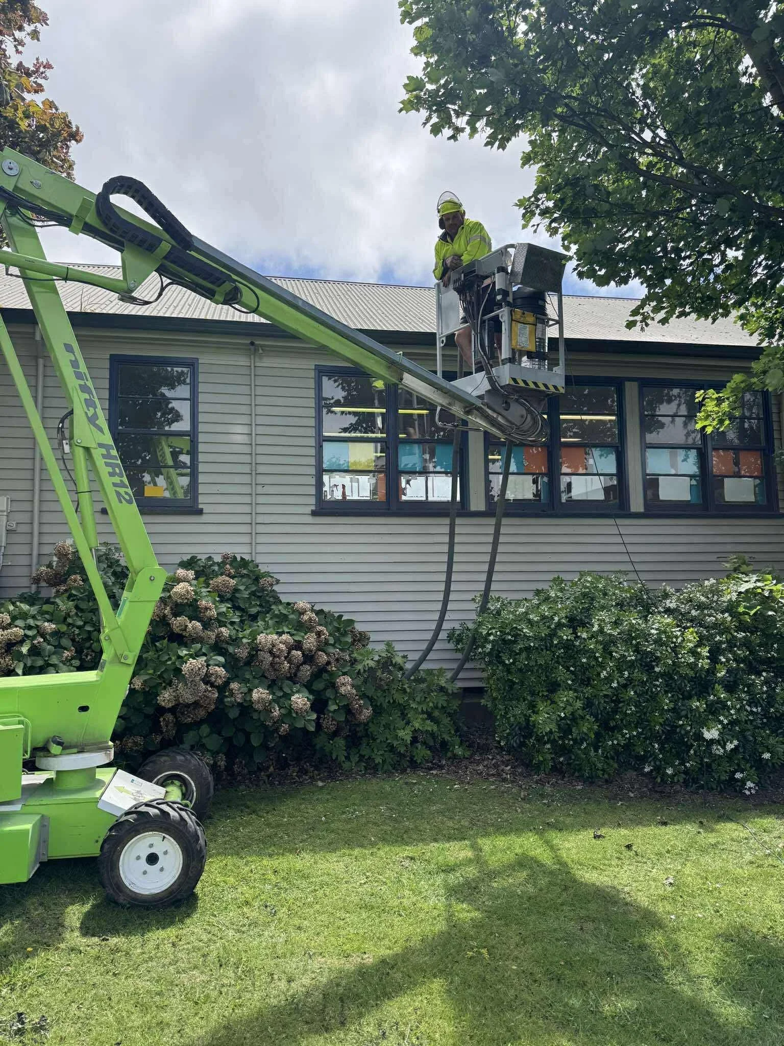 A property maintenance worker standing in a cherry picker, working on the roof of a building. The lift arm extends towards the building to clean the gutters and wash the building
