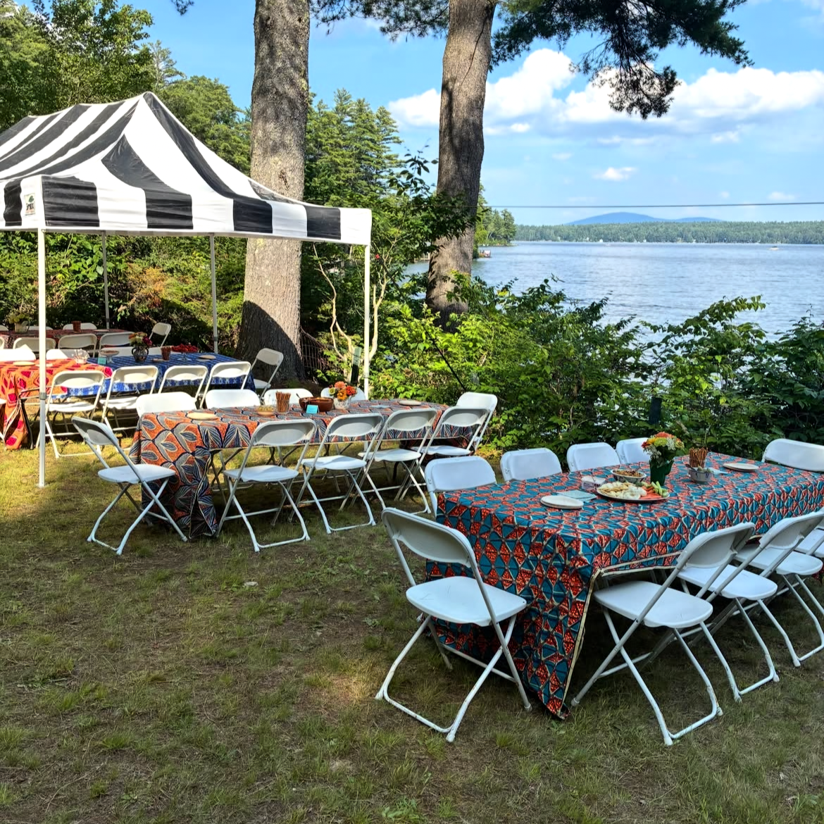 Outdoor lakeside party setup with tables covered in colorful tablecloths, surrounded by white chairs, under a striped canopy, near trees and water with a scenic view of the lake and sky in the background.