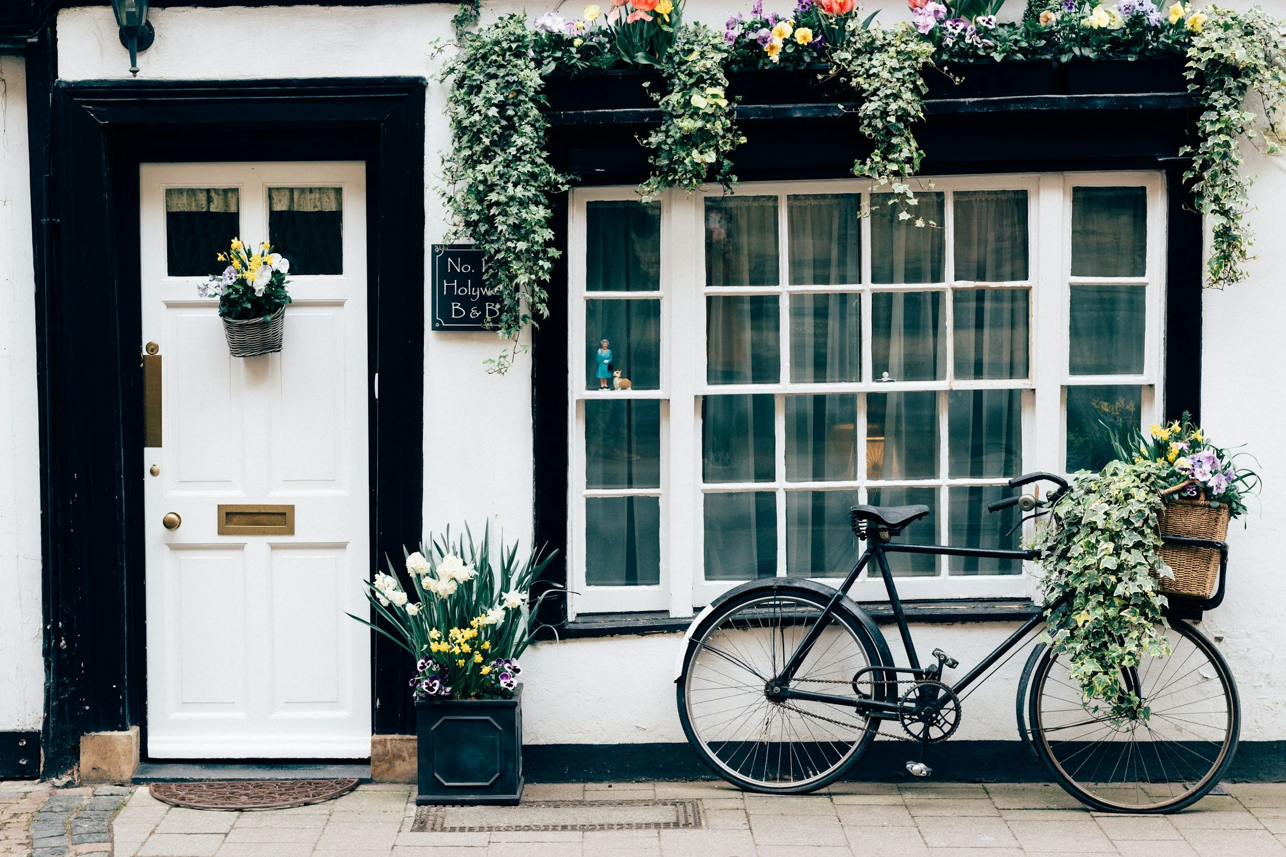 A white front door with a brass mail slot and a vase of colorful flowers hanging on the door handle. To the right, a large window with multiple panes, decorated with several potted plants and flowers. A black bicycle is parked against the wall below the window. There is a black signboard next to the door with white writing that says, "No. Holywah B & B". The ground is paved with bricks and stones.