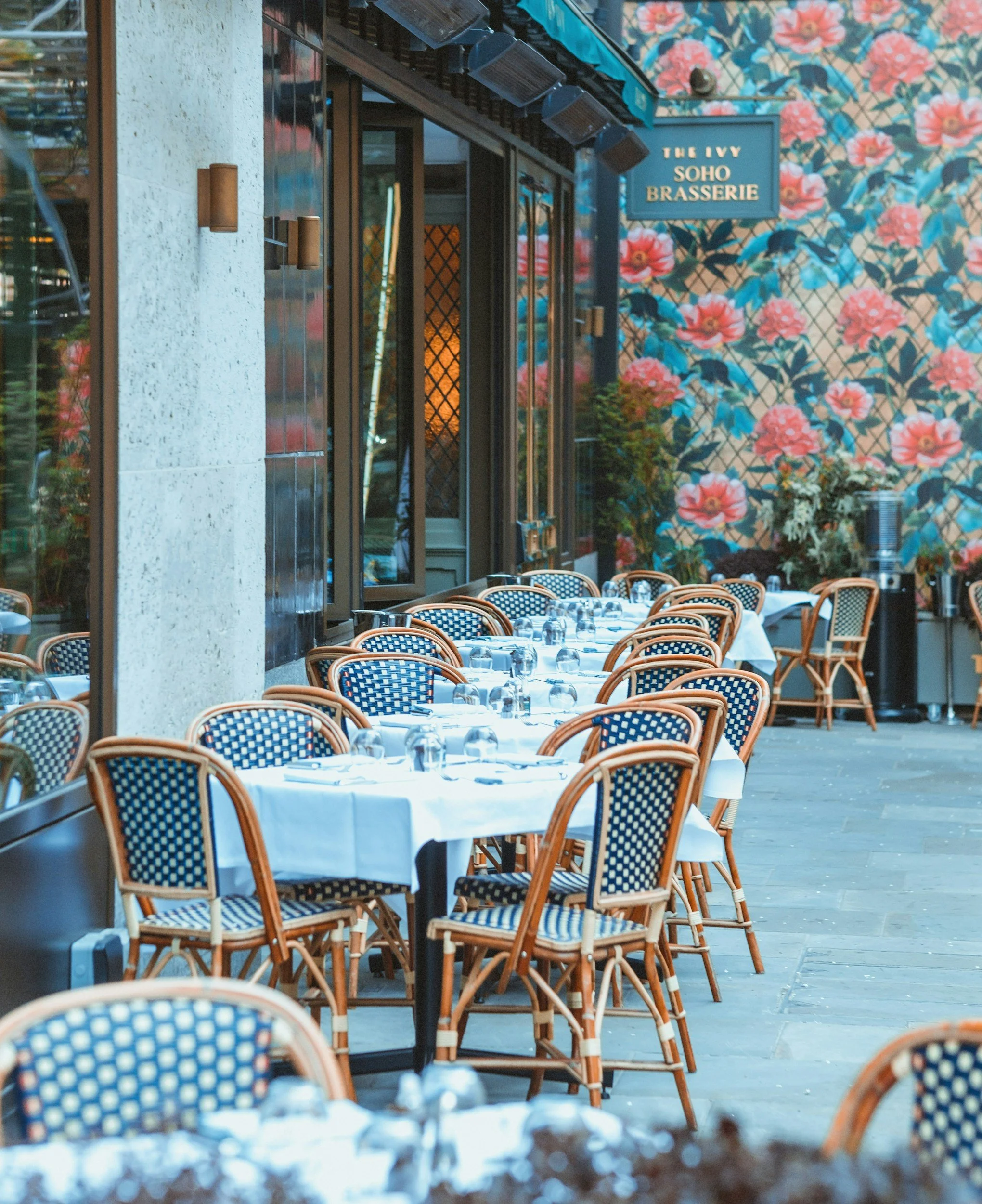 Empty outdoor terrace of a restaurant with white tablecloths, glasses, and chairs, situated near a wall with floral wallpaper and a sign that reads "The Ivy Soho Brasserie".