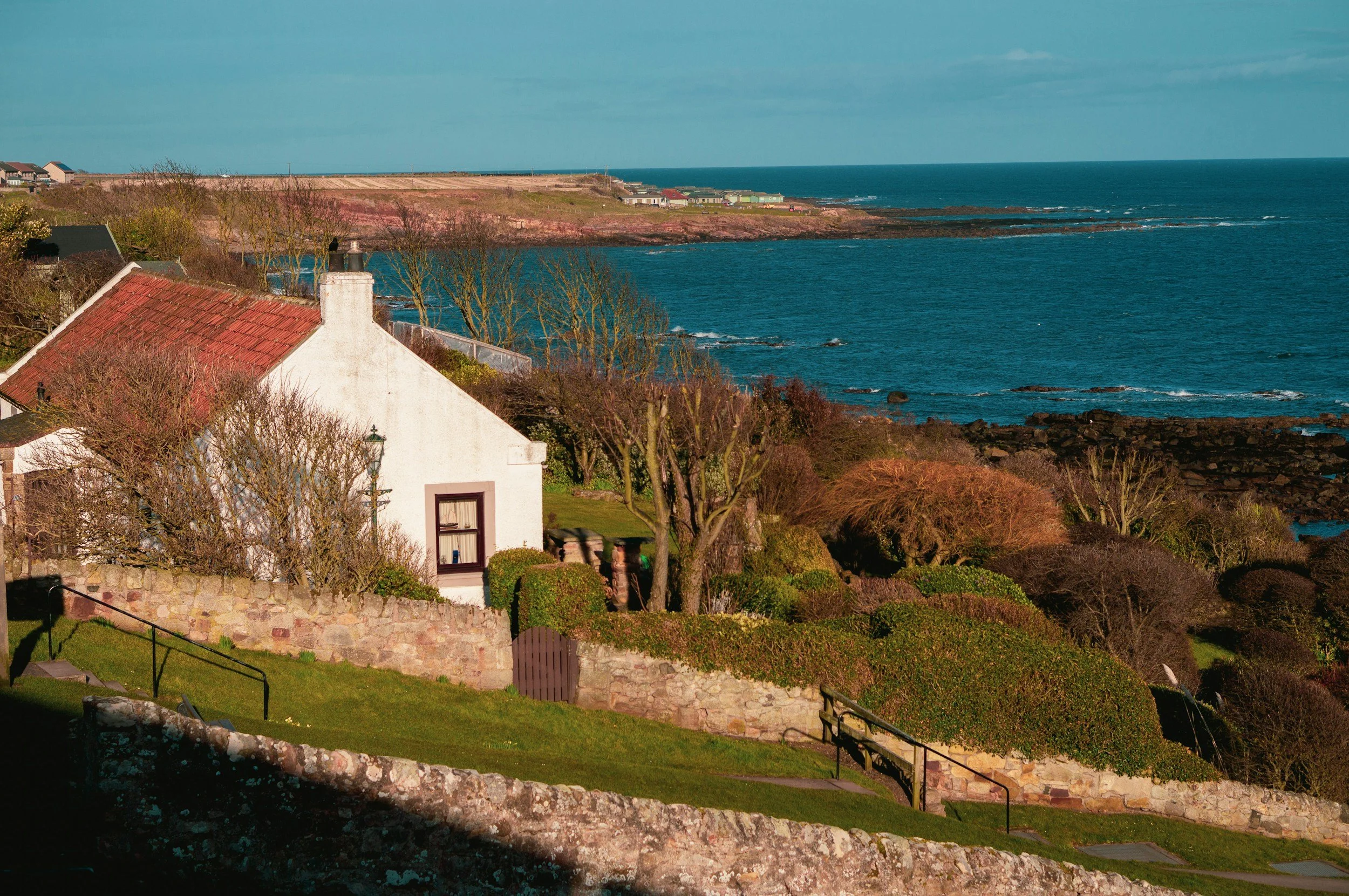 A coastal scene with houses, trees, and a stone wall in the foreground, overlooking the ocean and a sandy shoreline with cliffs in the background.