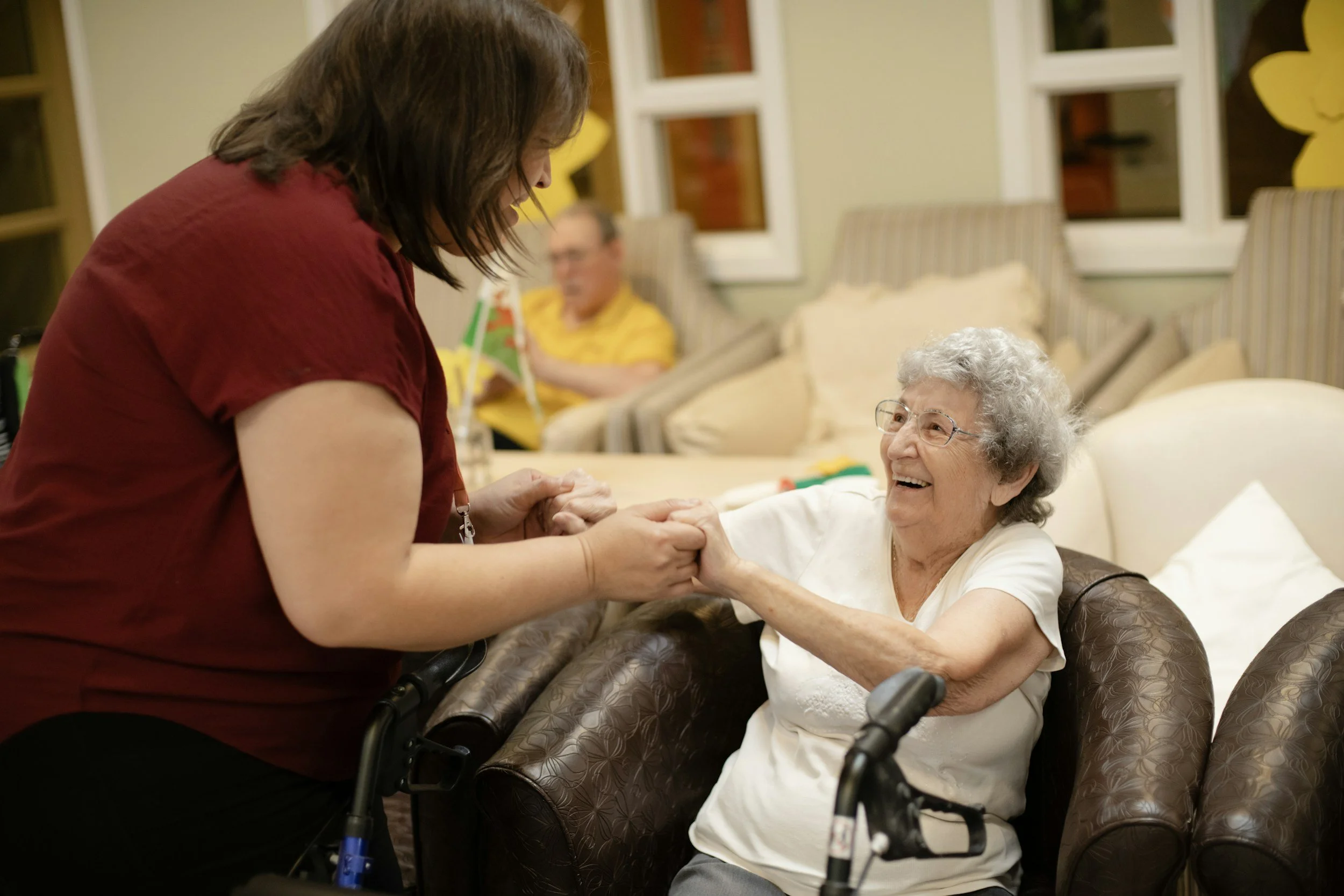 A woman in a red top is holding hands with an elderly woman seated in a wheelchair, smiling at her. In the background, a man in a yellow shirt is sitting on a couch, holding a small green and white object. The scene takes place in a cozy, well-lit room with windows decorated with yellow flower shaped cutouts.