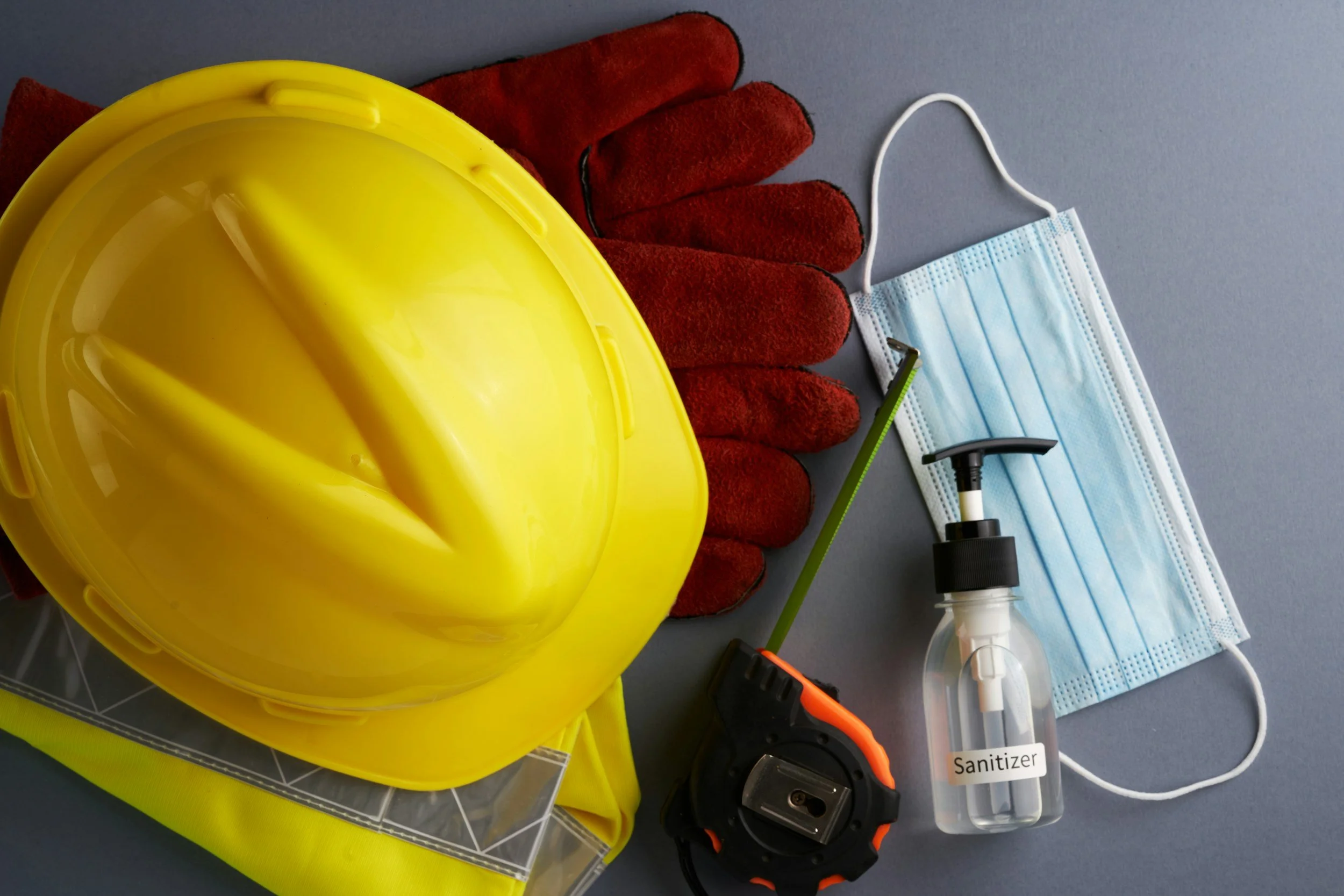 Safety equipment including a yellow hard hat, red work gloves, a face mask, a sanitizer spray, and other cleaning supplies on a gray surface.