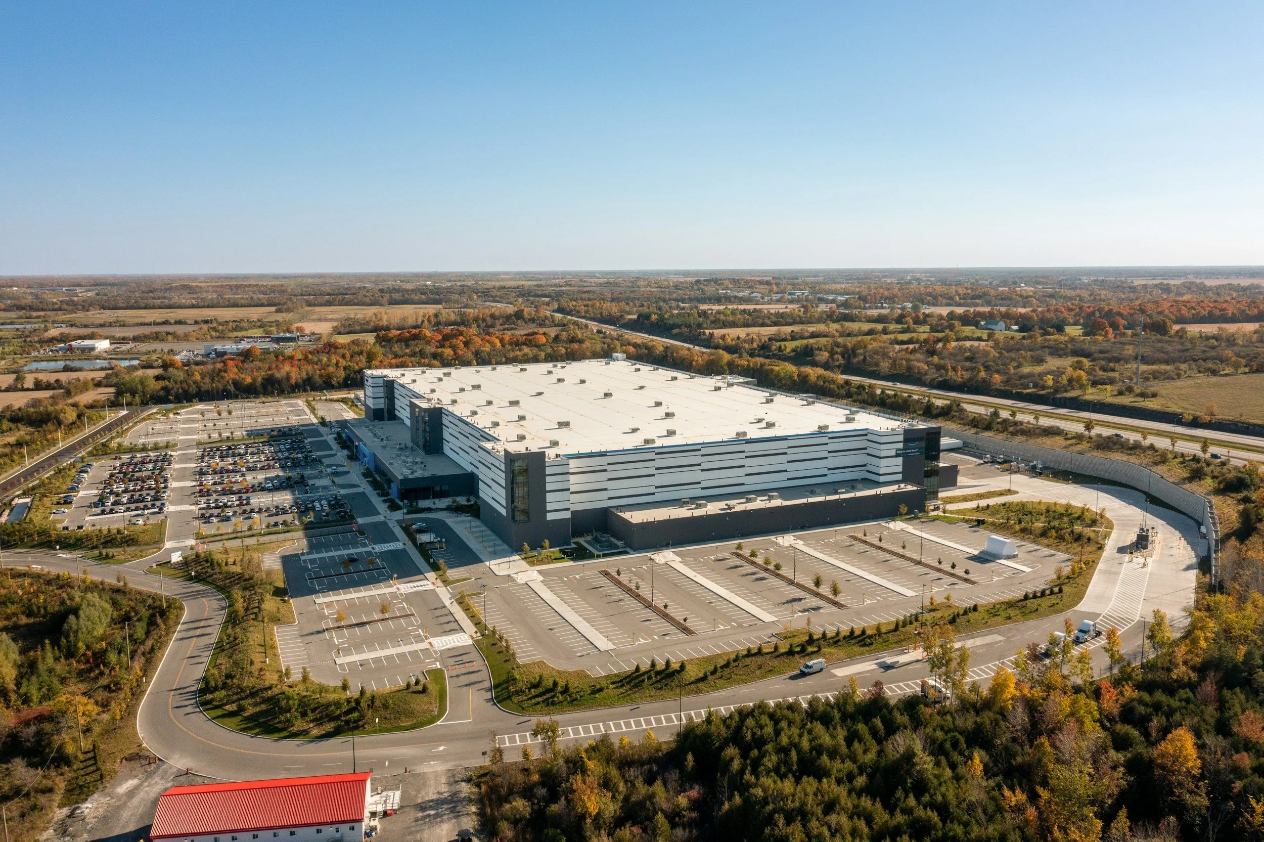 An aerial view of a large white commercial building with a parking lot, surrounded by trees and open fields, under a clear blue sky.