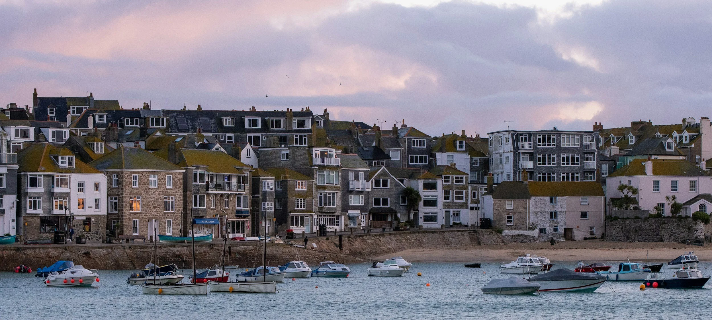 A coastal scene with boats docked in calm water and a hillside filled with tightly packed houses with various architectural styles and colors, under a cloudy sky during dusk.