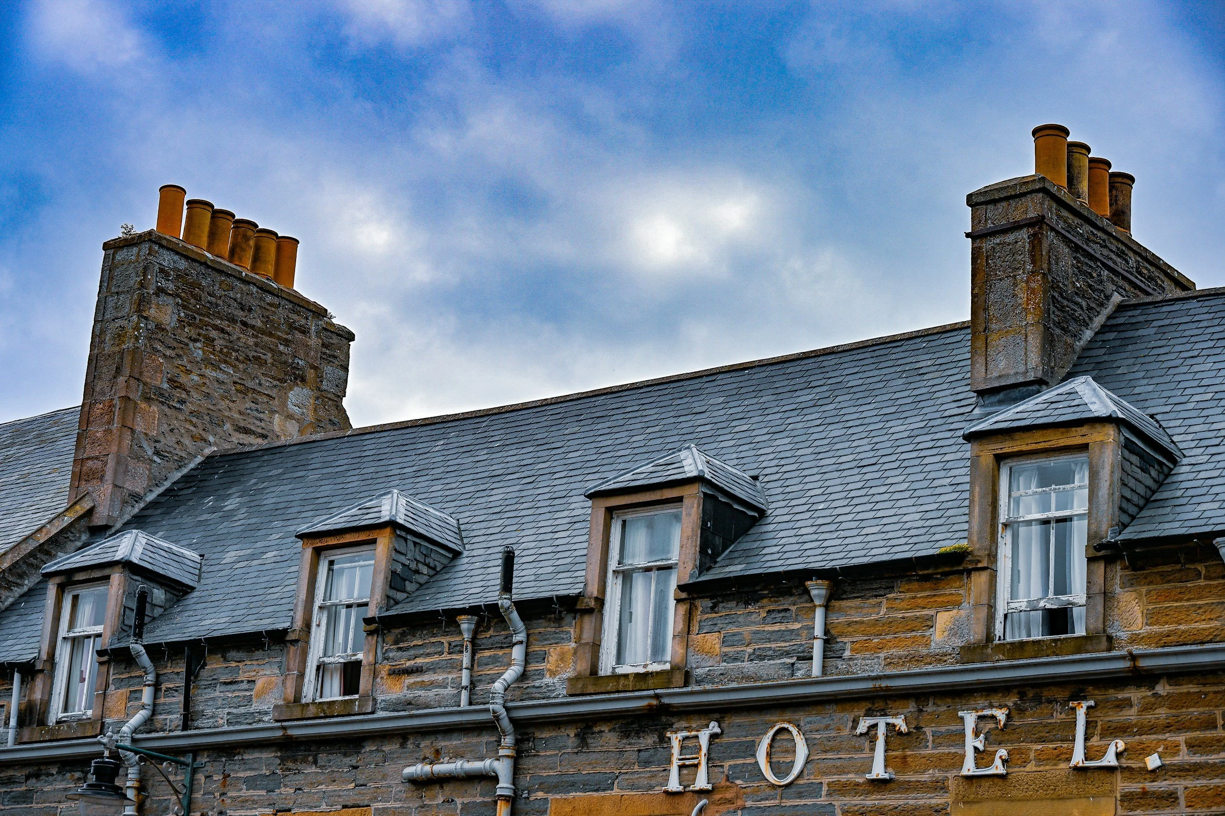 Close-up of a house with a stone brick exterior, slate roof, multiple dormer windows, and two brick chimneys with multiple yellowish chimney pots against a cloudy sky.