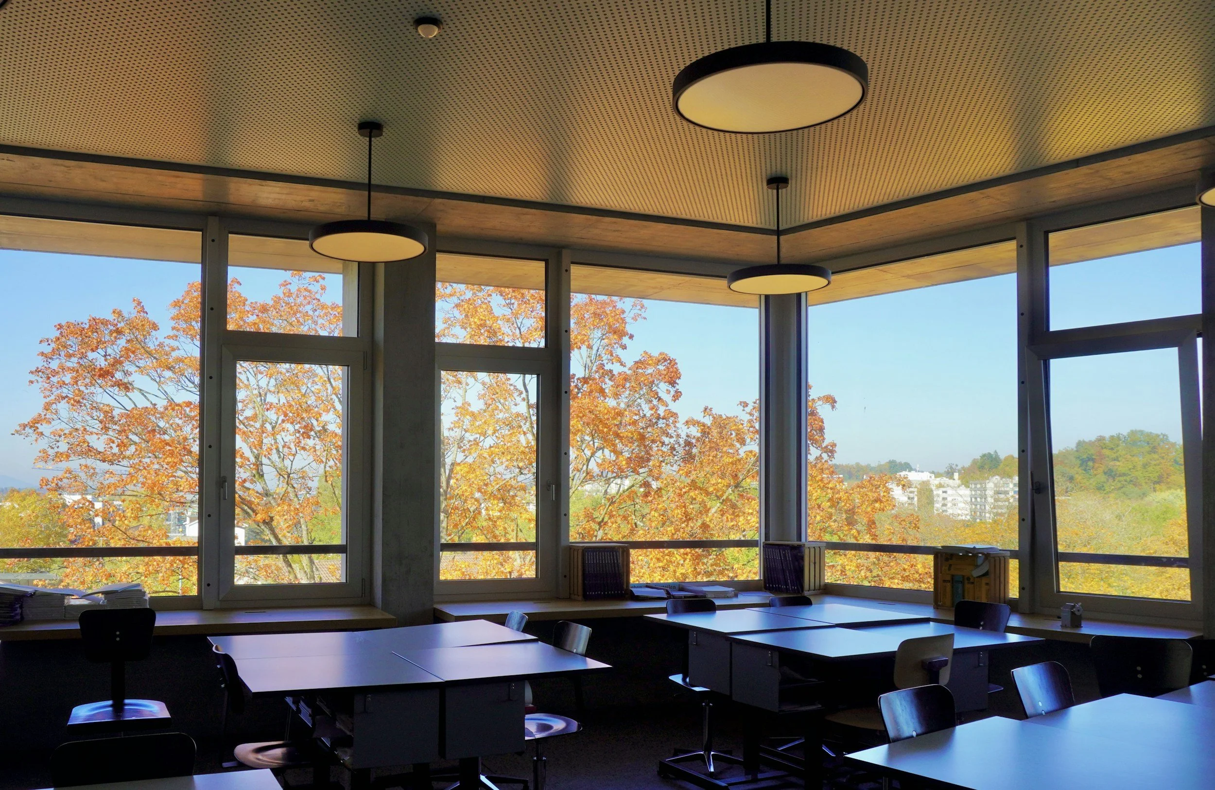 An interior classroom or conference room with large windows showing trees with autumn foliage. The room has modern ceiling lights and tables with chairs, some papers and books on the tables.