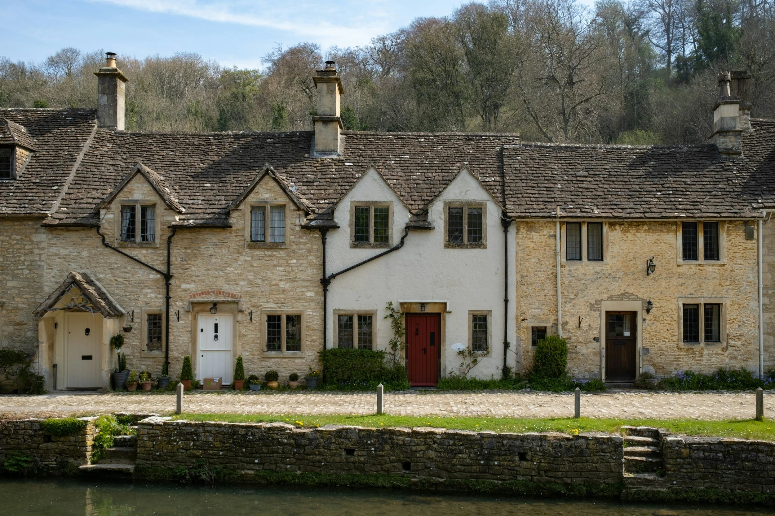 A row of historic stone cottages with sloped shingled roofs, small windows, and individual doors next to a canal or river, with trees and clear sky in the background.
