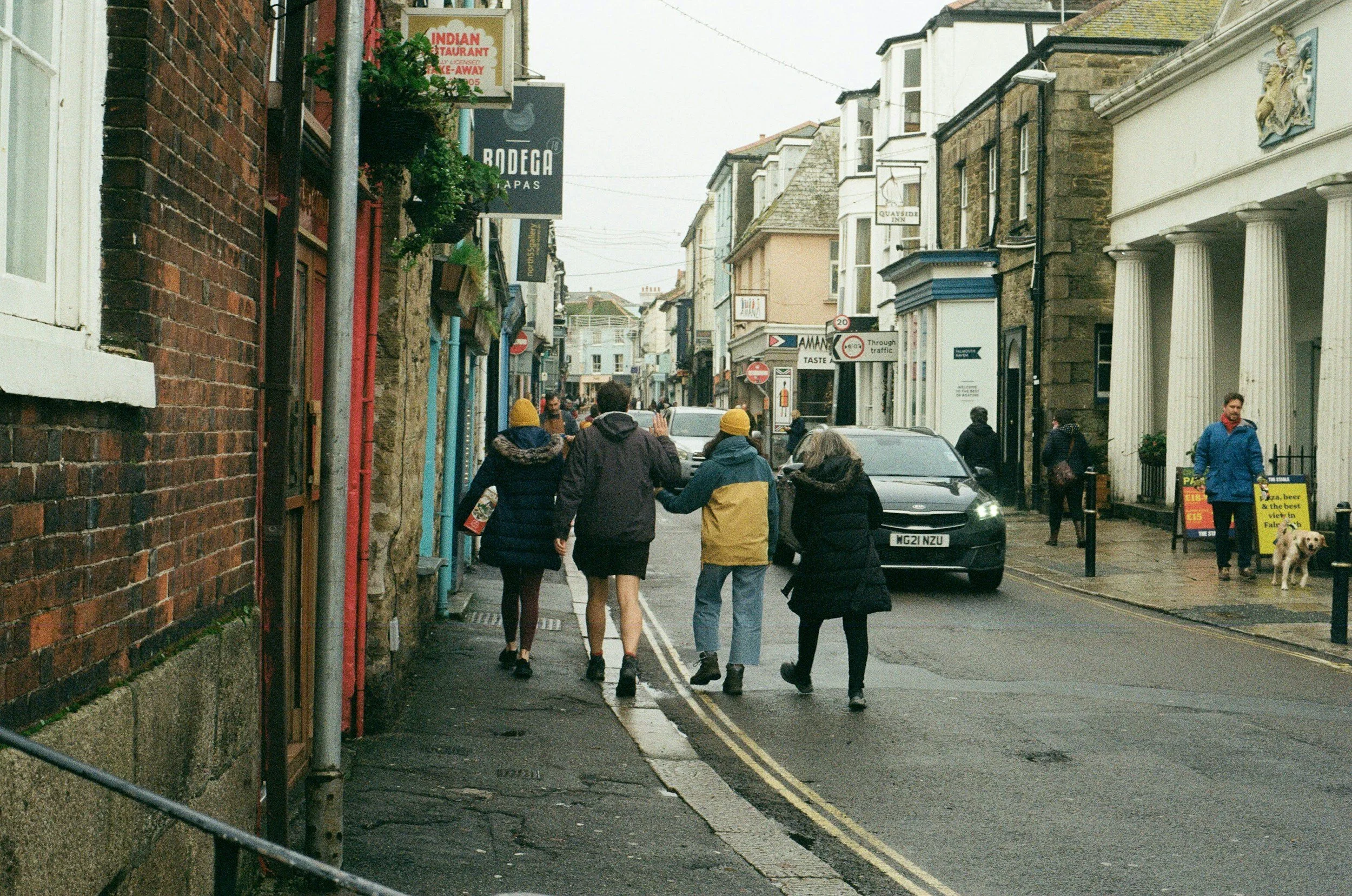 A busy street scene with pedestrians walking on the sidewalk and cars parked along the curb in a town with various shops and signs, including an Indian restaurant and a tapas bar.