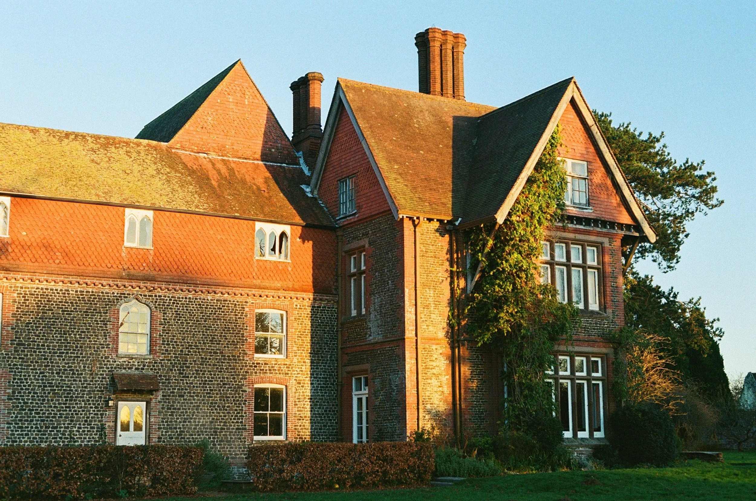 A historic brick and stone building with multiple gabled roofs, chimney stacks, and arched windows, illuminated by warm sunlight against a clear blue sky.