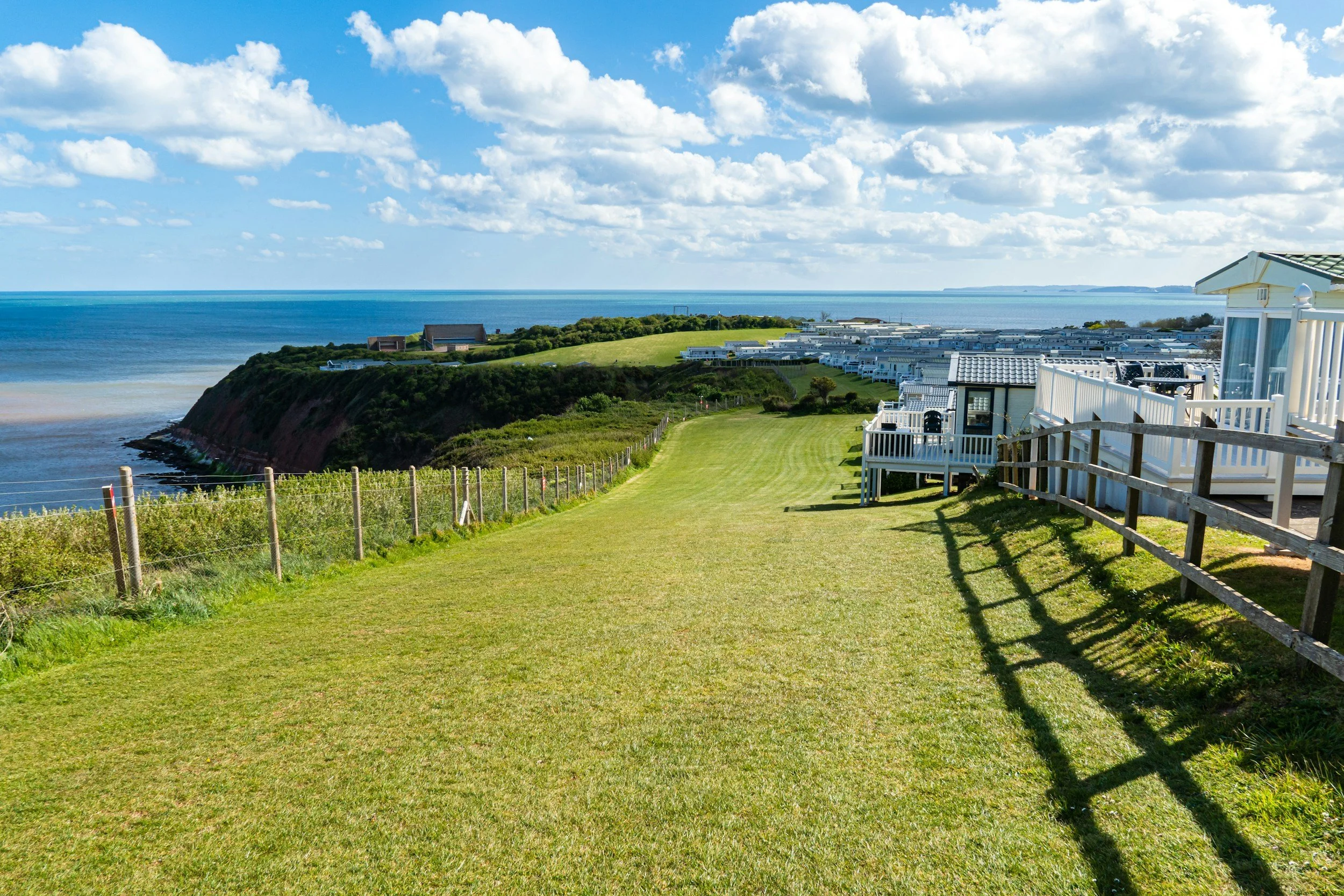 Scenic view of a grassy hill with houses and fencing, overlooking the ocean and a partly cloudy sky.