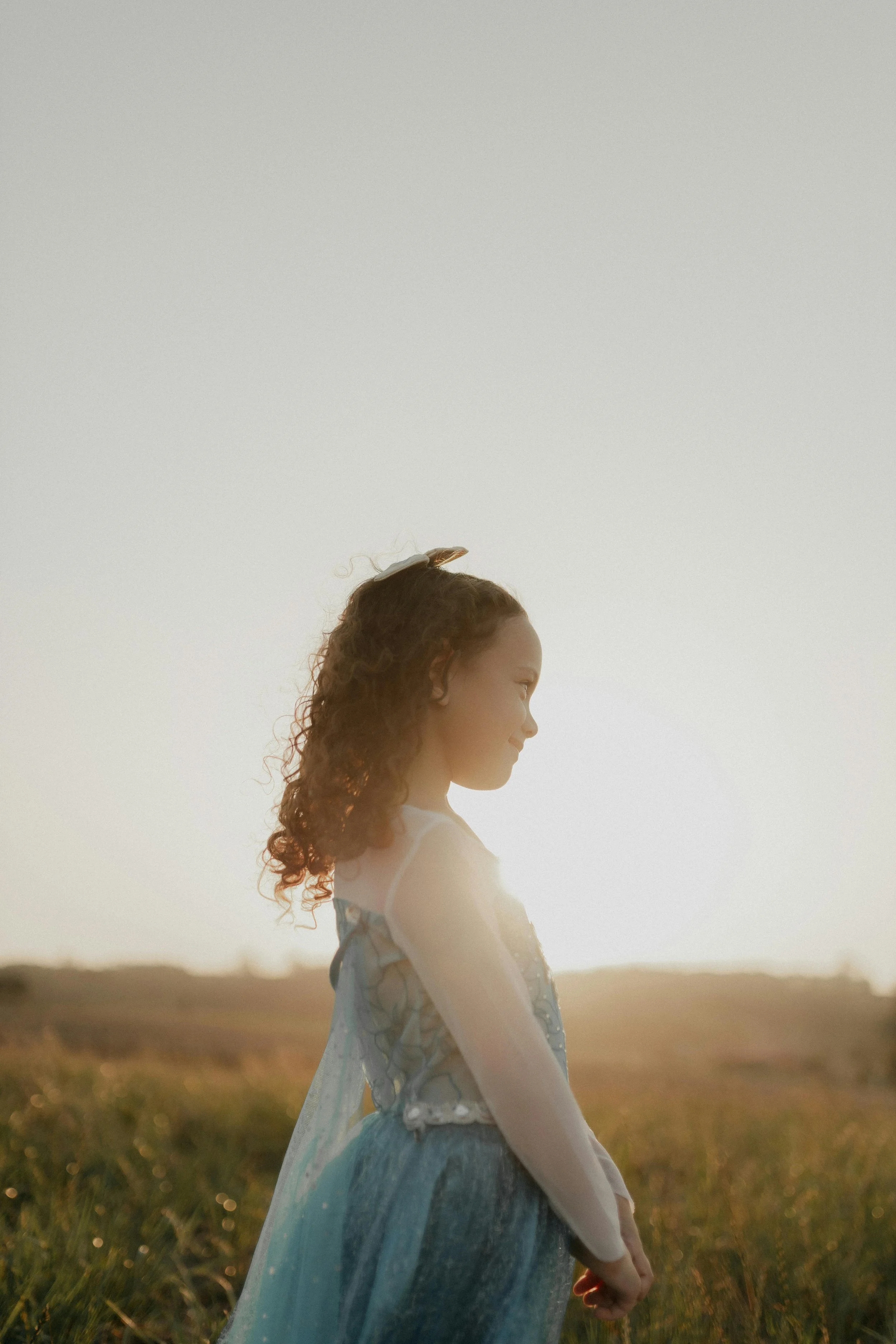A young girl with curly hair standing in a field during sunset, wearing a light blue dress with floral patterns, facing sideways with a peaceful expression.