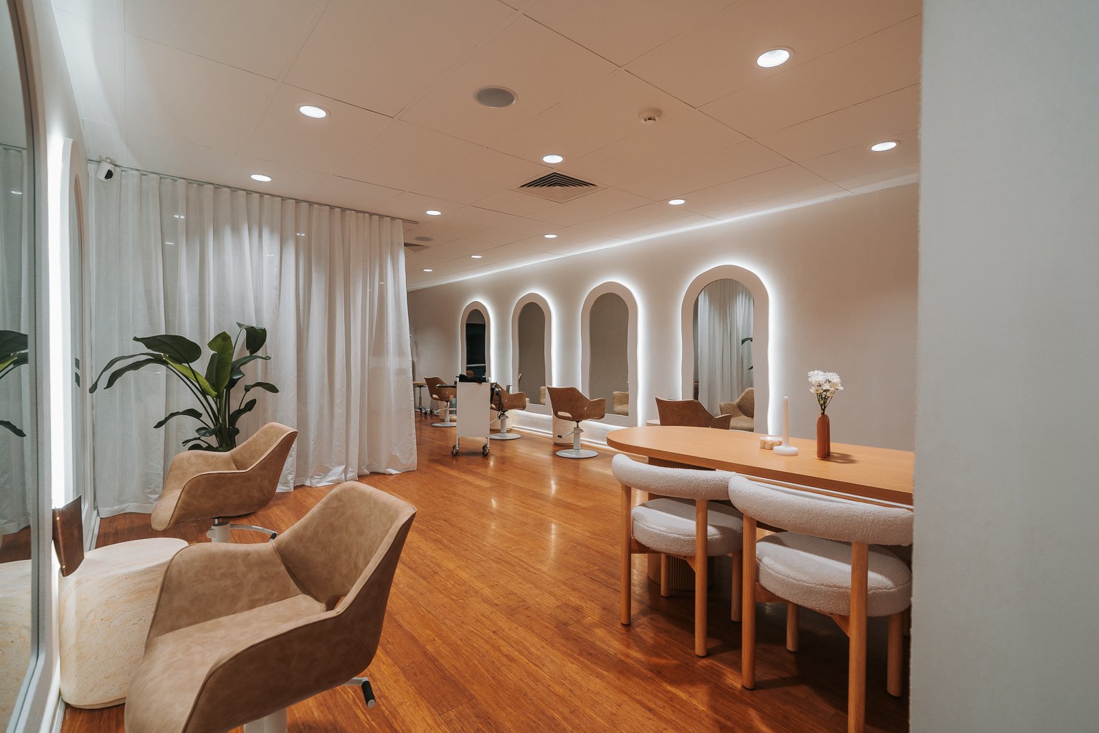 Modern salon with wooden floors, beige chairs, white curtains, and illuminated mirrors on the wall.